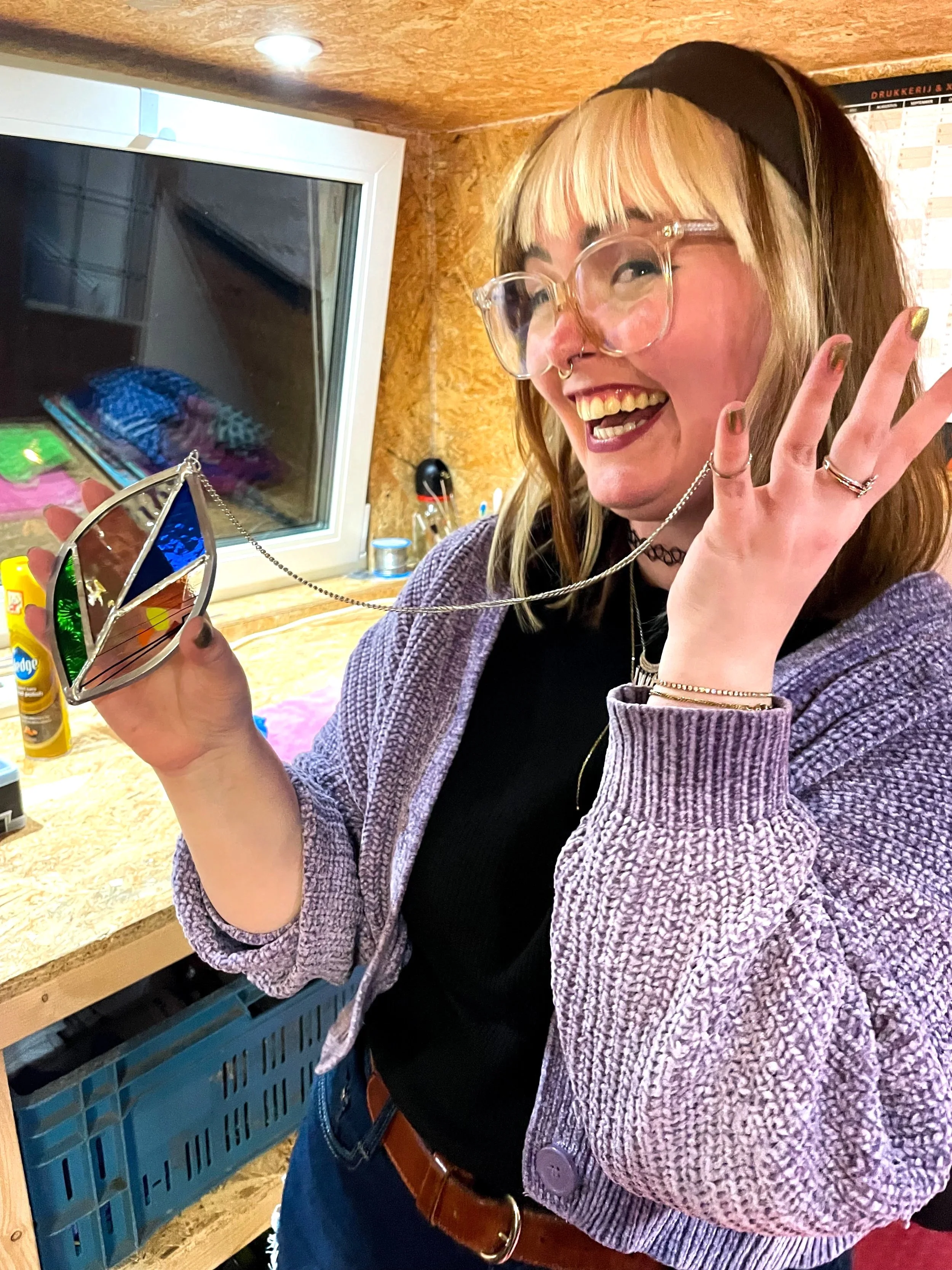 Een vrouw met blond haar en een zwart haarbandje, lachend en gekleed in een zwarte trui en een paarse jas, houdt een spiegel met een kleurrijk frame in haar hand. Ze staat binnen in een kamer met houten muren en een raam achter haar.