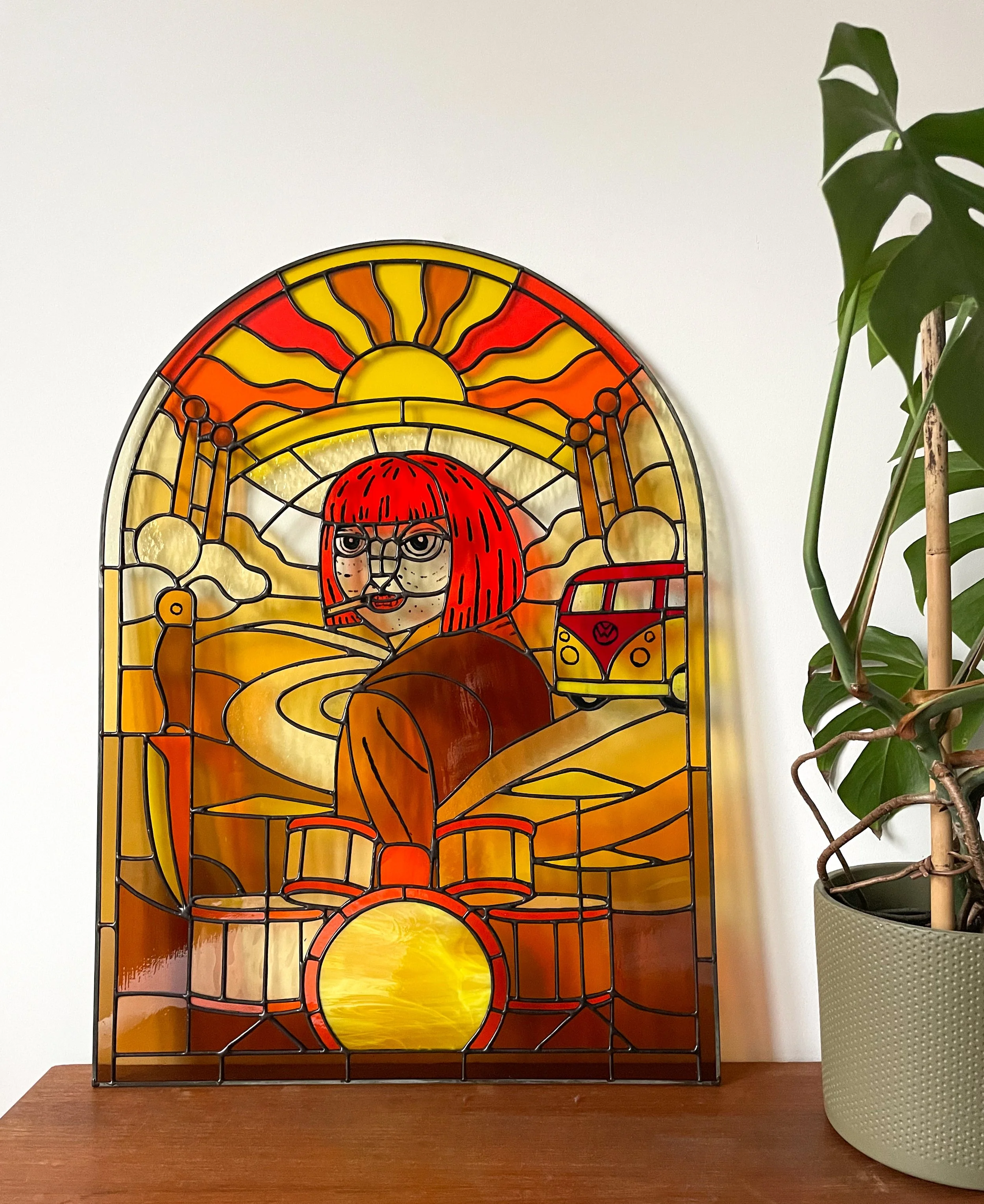 Kleurriek glas-in-lood raam met een zon en een persoon met rode haar en bril, naast een groene plant in een witte pot.