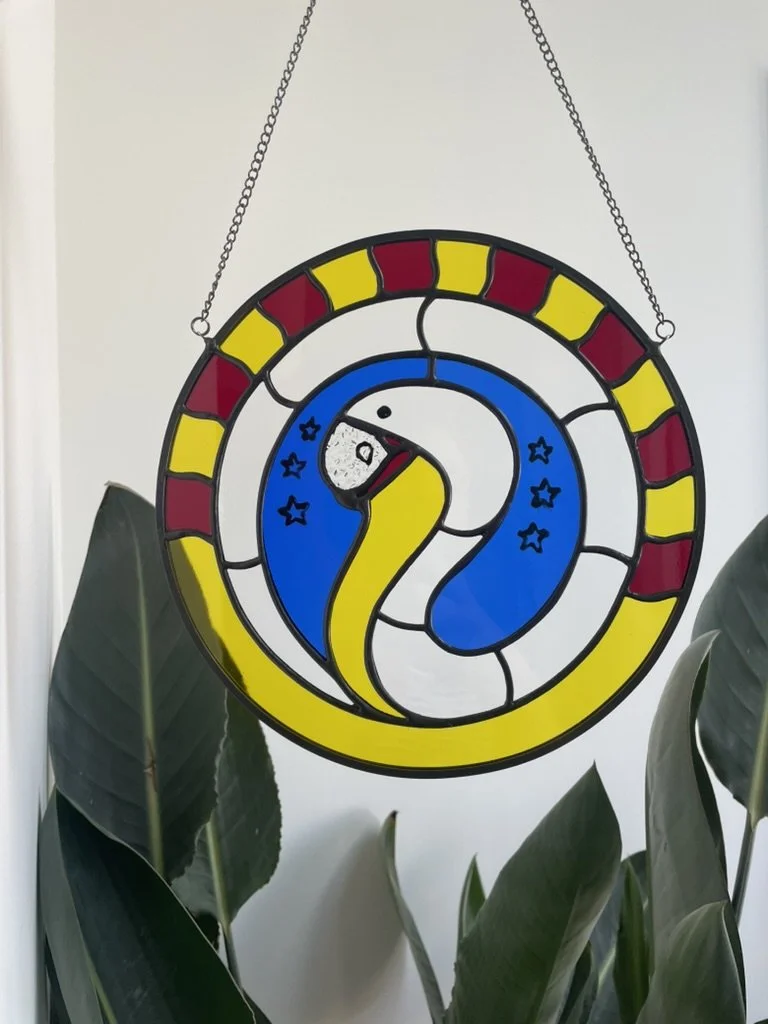 Kunstwerk naar de alfbeelding van Daniel Romano in glas met de afbeelding van een slang met een veelkleurige achtergrond, boven een plant met grote groene bladeren.