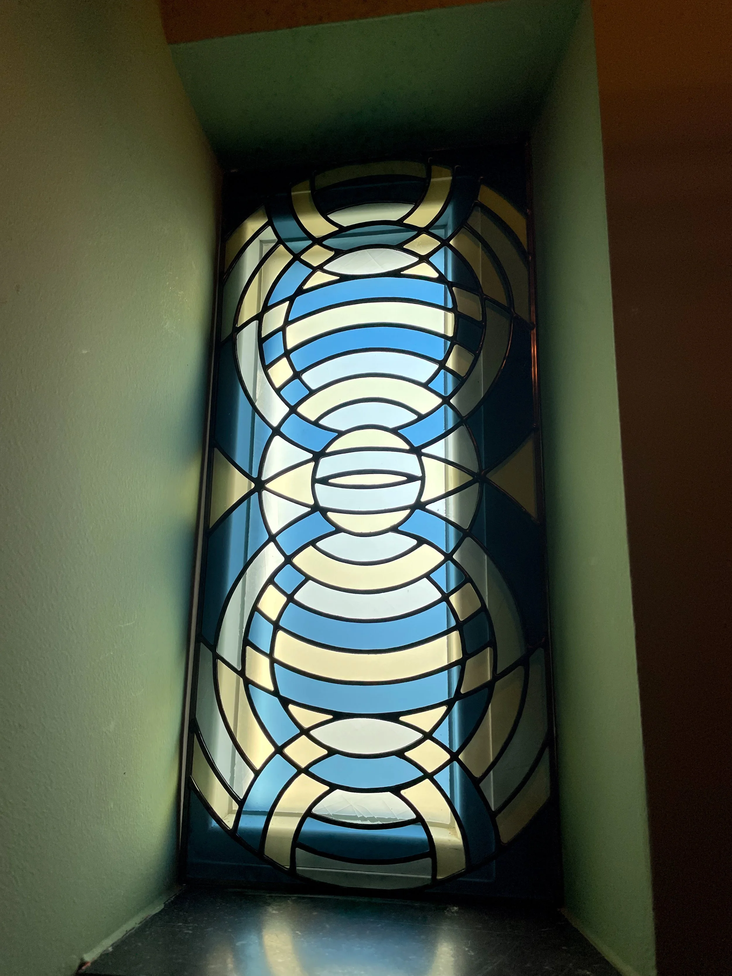 Glas-in-lood raam met abstract patroon en blauwe, gele en witte kleuren, geplaatst in een nis tussen groene muren.