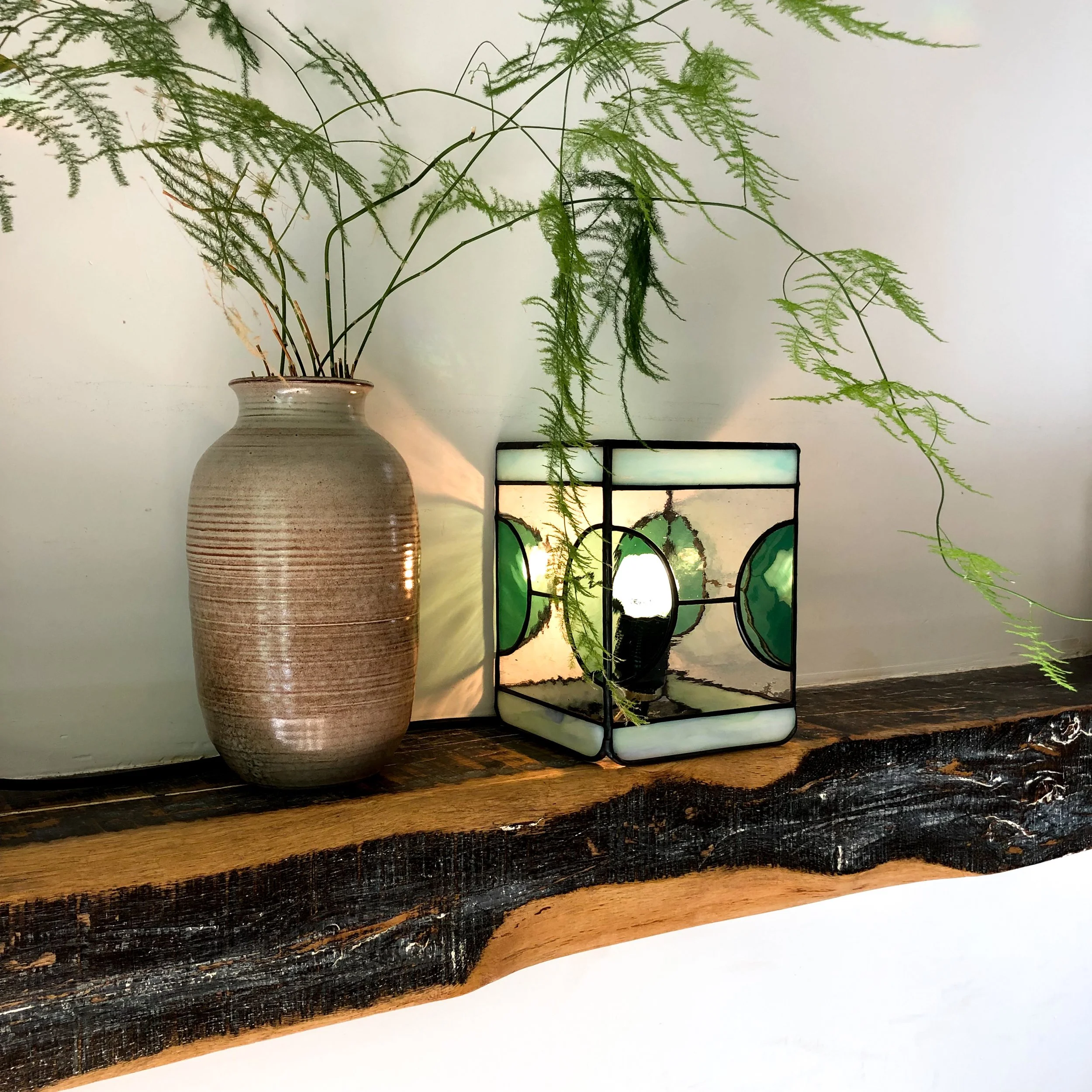 Groene planten in een keramische vaas, geplaatst op een houten oppervlak, met een decoratief lantaarn van glas erachter.