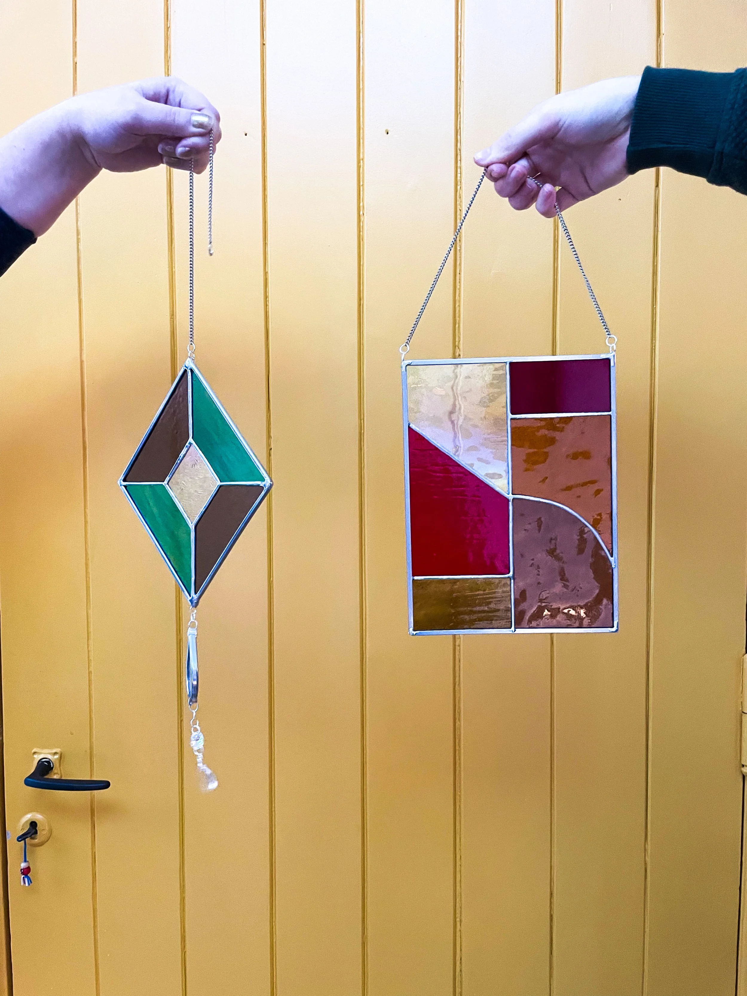 Twee handen houden gekleurde glazen kunstwerken, een ornament met een geometrisch patroon en een ander met een abstract ontwerp, hangend aan de deur.