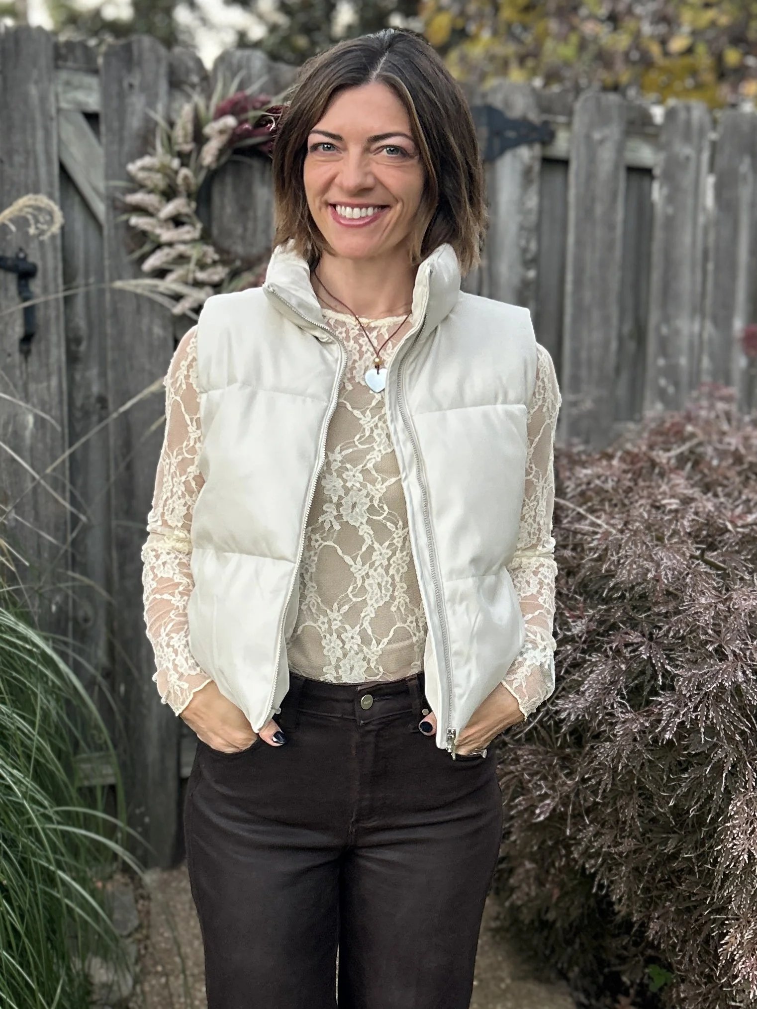 Woman with short brown hair smiling outdoors in a garden, wearing a light-colored vest over a lace top and dark pants.