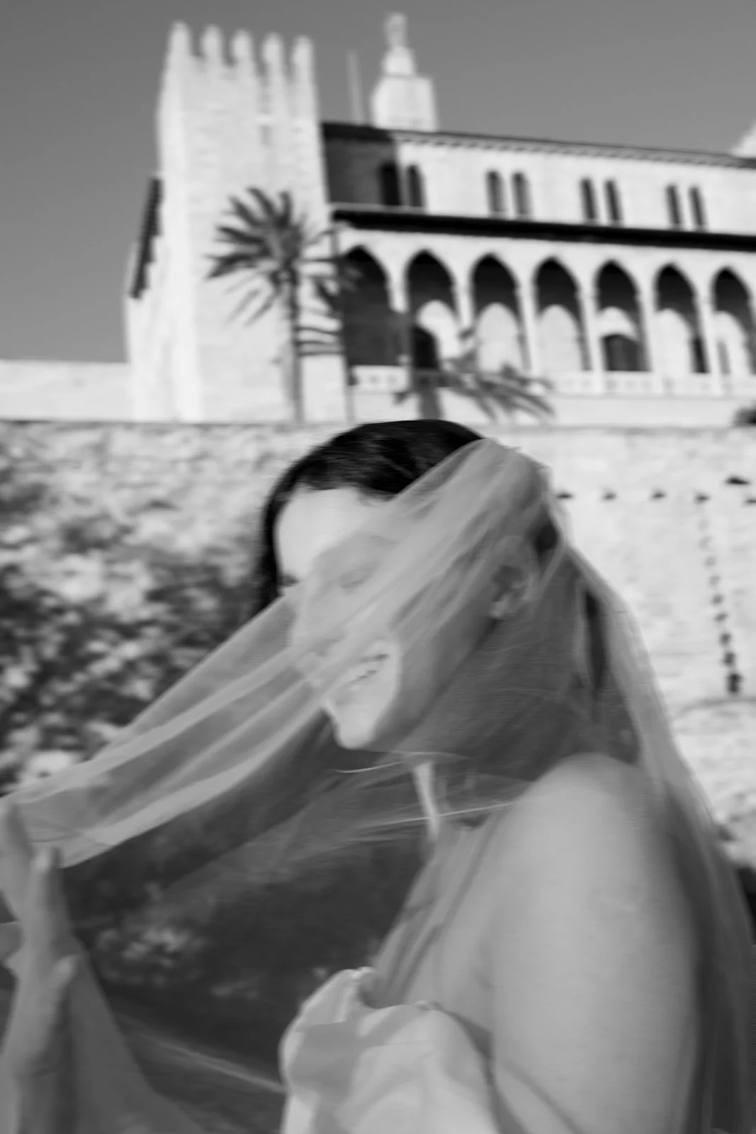 A woman with dark hair, wearing a face shield, in front of a large building with a tower, arches, and a palm tree in the background.