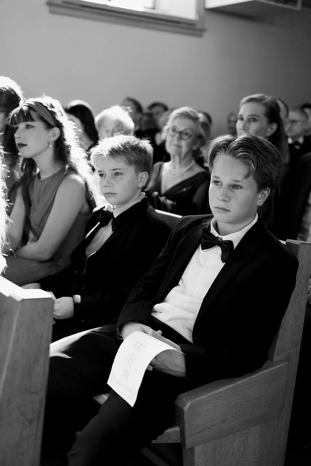 People attending a formal event, seated in a church or auditorium, dressed in suits and dresses, watching an ongoing ceremony or presentation in black and white.