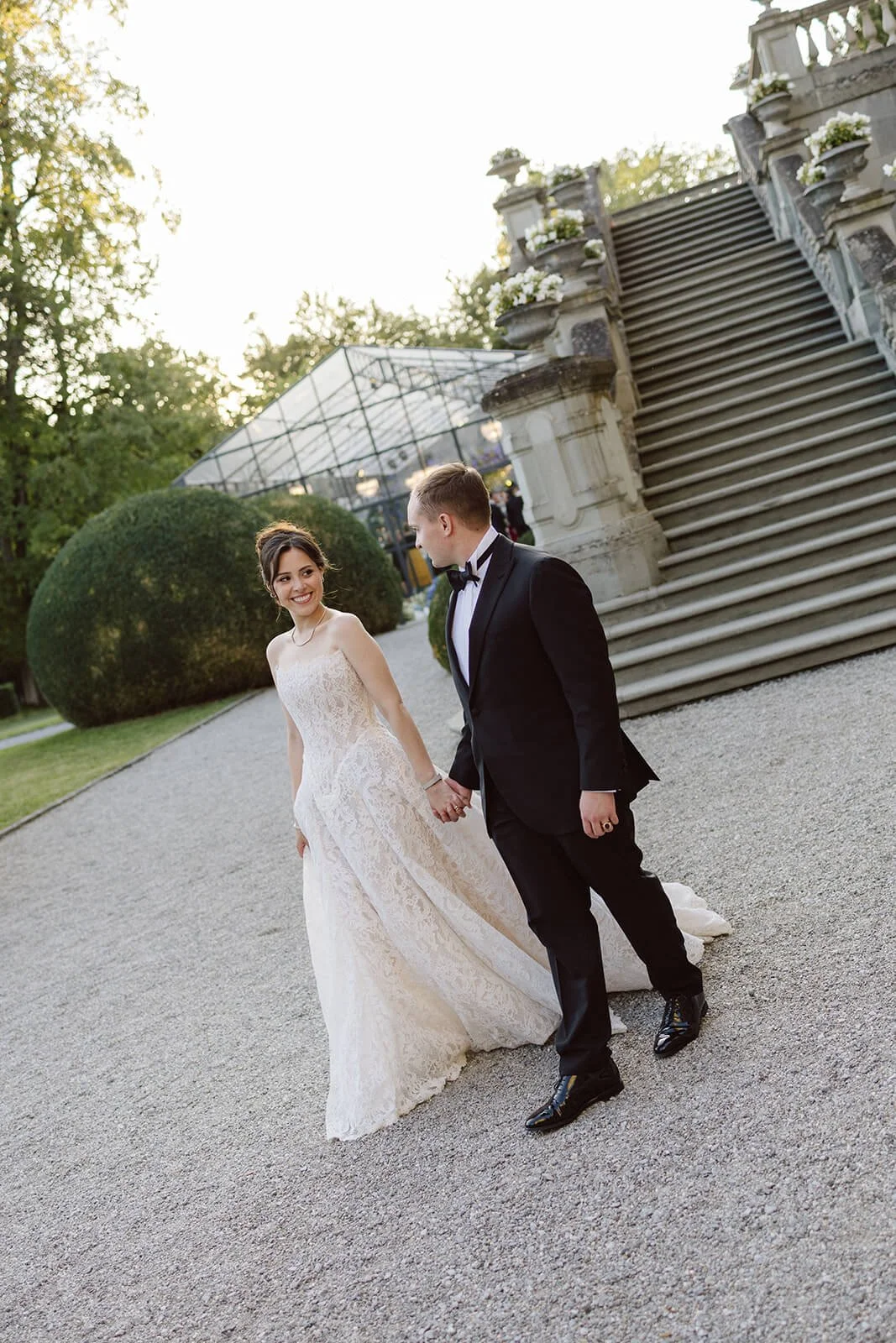 A bride and groom holding hands and smiling at each other outdoors near stone stairs and large bushes on a gravel pathway, with sunlit trees in the background.