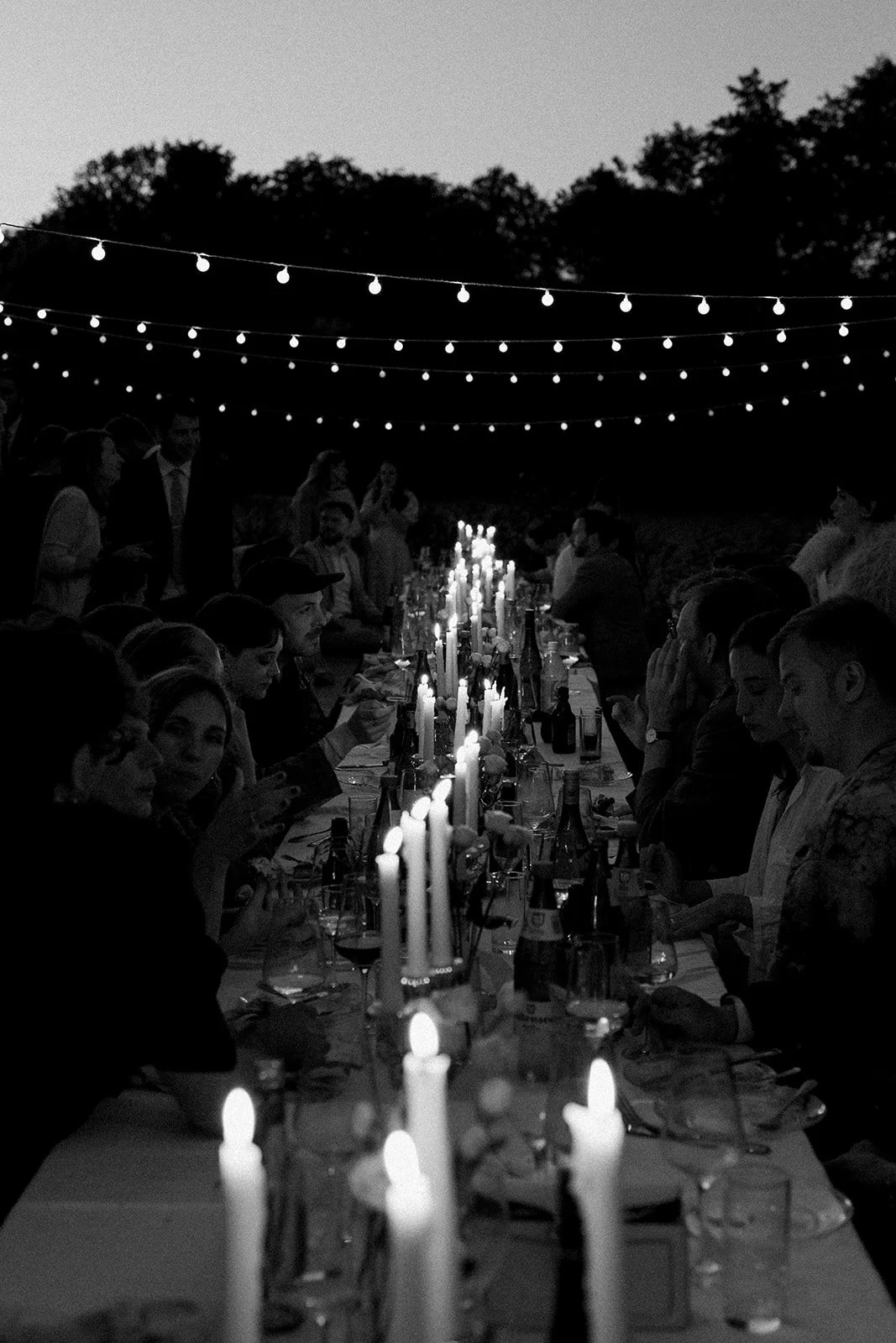 A black-and-white photo of an outdoor dinner party at dusk, with a long table decorated with lit candles and surrounded by guests, under string lights and trees in the background.