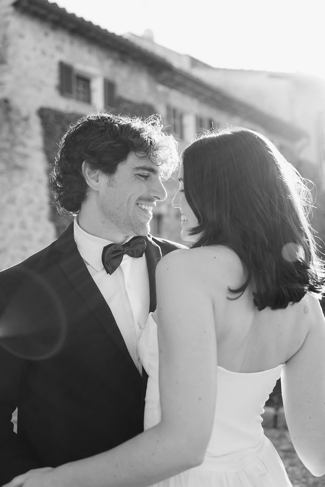 A black and white photo of a smiling couple standing outdoors, close together, with a rustic building in the background. The man is wearing a tuxedo with a bow tie, and the woman is in a strapless dress with shoulder-length hair.