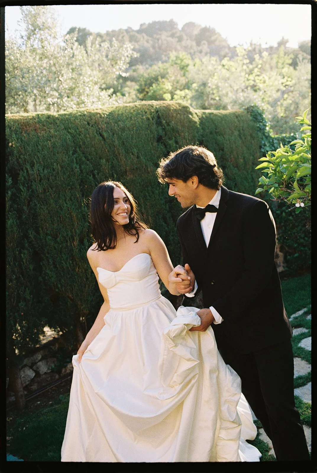 A woman in a white strapless wedding dress and a man in a black tuxedo hold hands and smile at each other outdoors, with greenery and trees in the background.