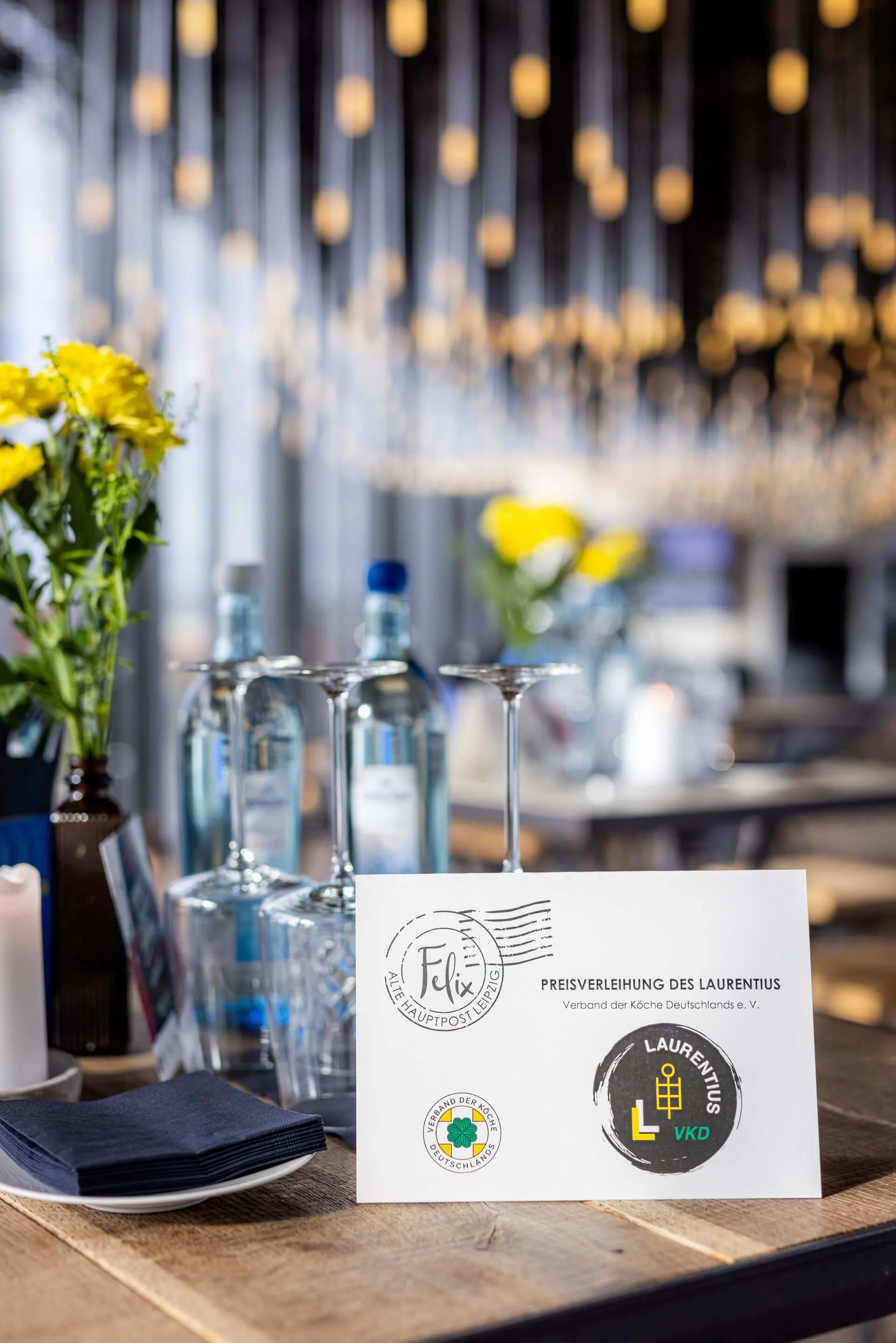 A table with a certificate, black napkins, and two water bottles inside a restaurant or event space with hanging light bulbs and blurred background.
