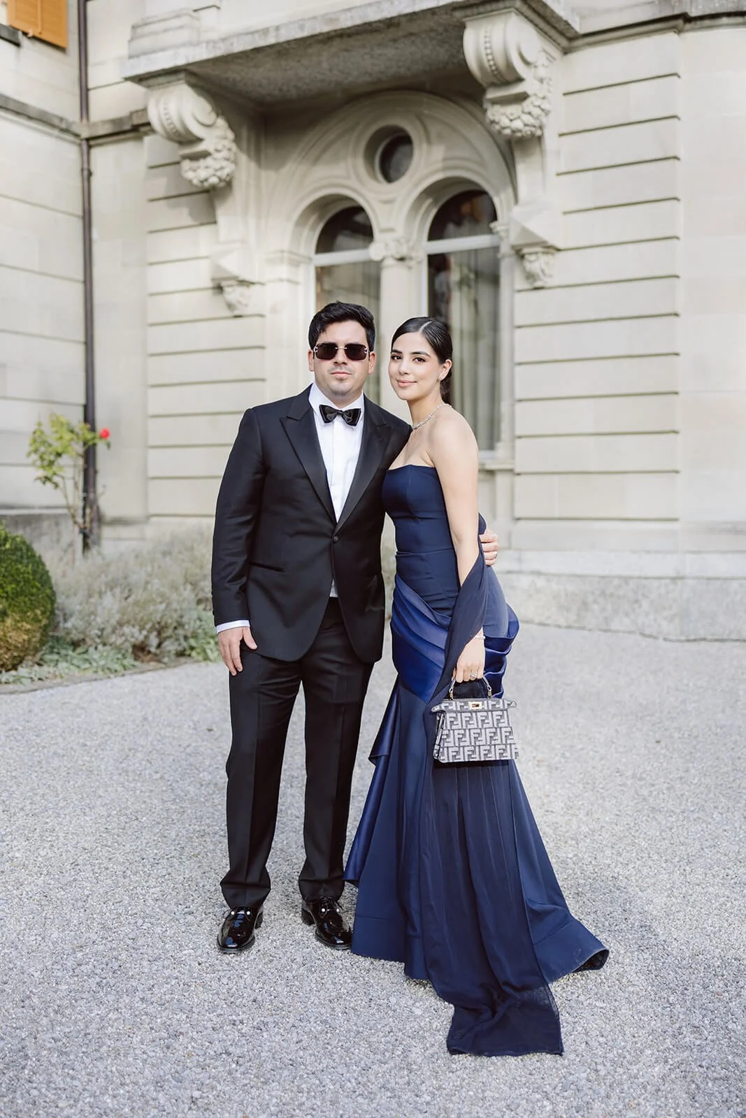 A man and woman dressed in formal evening attire standing outdoors in front of a historic building. The man is wearing a black tuxedo with a bow tie and sunglasses, while the woman is wearing a strapless navy blue gown and holding a designer handbag.