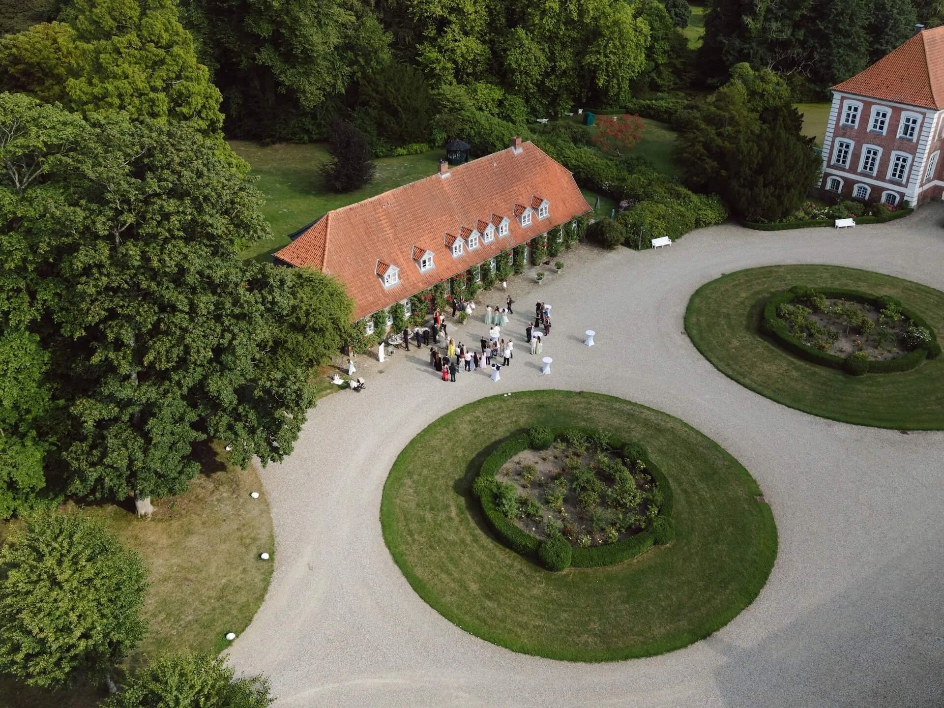 An aerial view of a wedding reception outdoors at a historic estate, with guests mingling near a long building with a red-tiled roof, surrounded by lush green trees, gardens, and curved pathways.