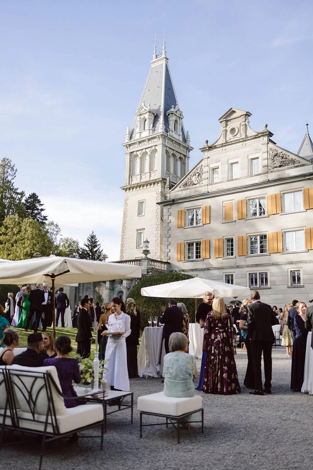 People gathered outdoors at a wedding reception or celebration in front of a historic castle with tall towers, beige walls, and yellow shutters. White umbrellas provide shade.