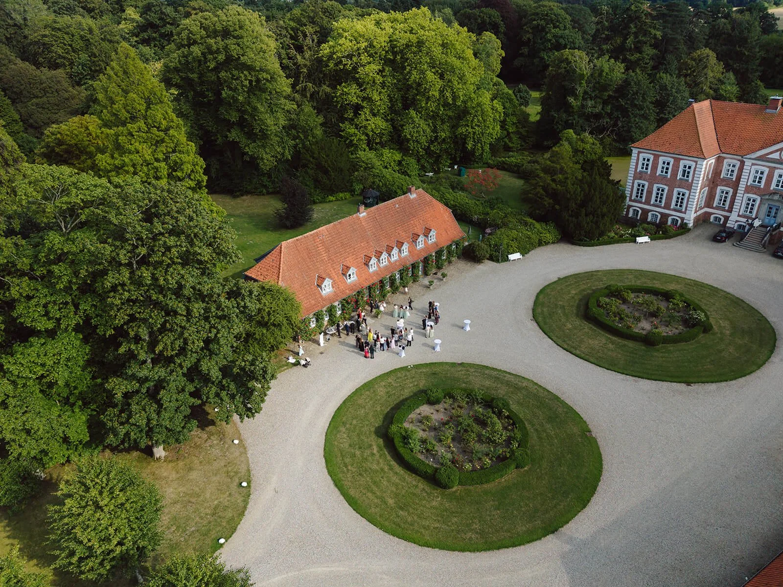 An aerial view of a large estate with a circular driveway, manicured gardens, and two prominent red-roofed buildings surrounded by lush trees, with people gathered near one of the buildings.