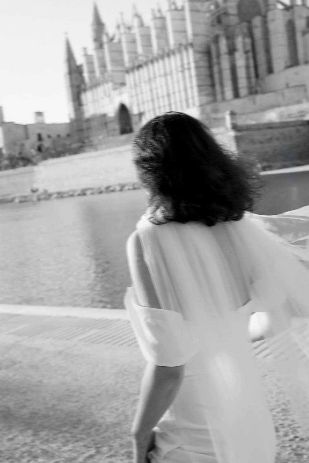 A woman with dark, curly hair tilts her head back, sitting on a bench by the water. In the background, a large historic building with turrets and arched windows is visible.