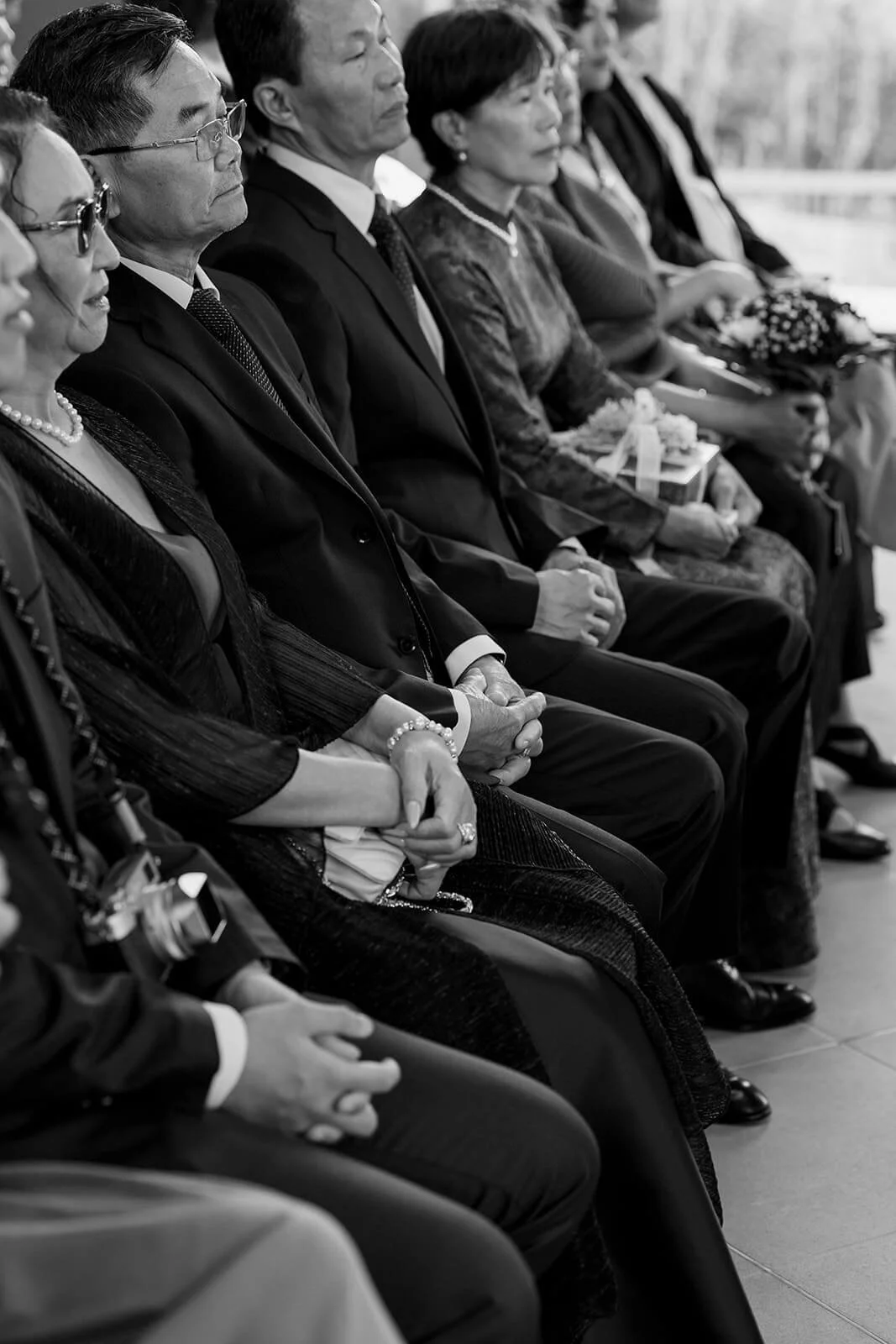 Black-and-white photo of a row of formally dressed people sitting with hands clasped, attending a solemn event or ceremony outdoors.
