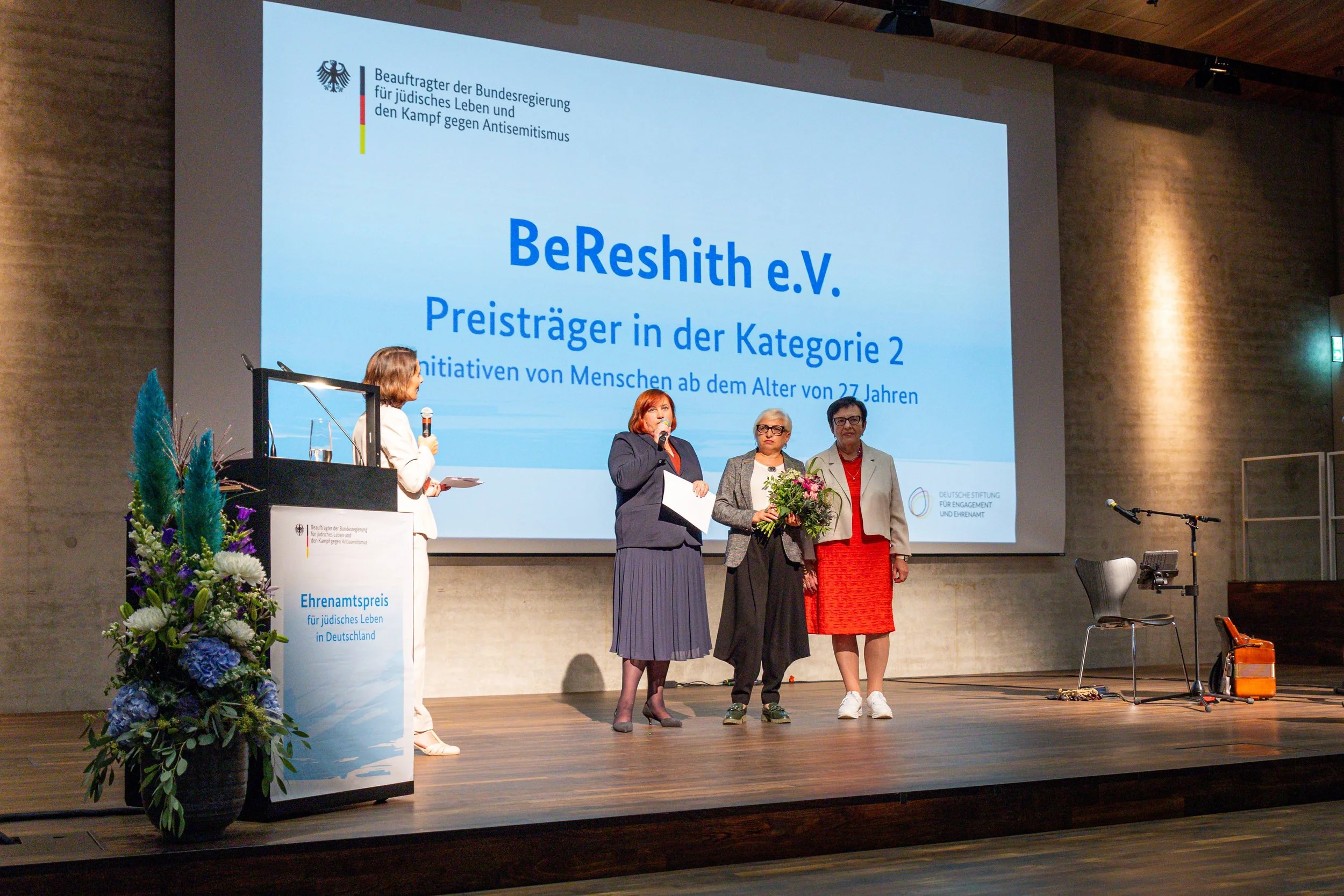 Women on stage at an award ceremony, with a large screen displaying text about the BeResith e.V. organization in German, including a banner with the German eagle emblem and the words 'Bundesregierung' and 'Antisemitismus.' One woman is holding a bouq