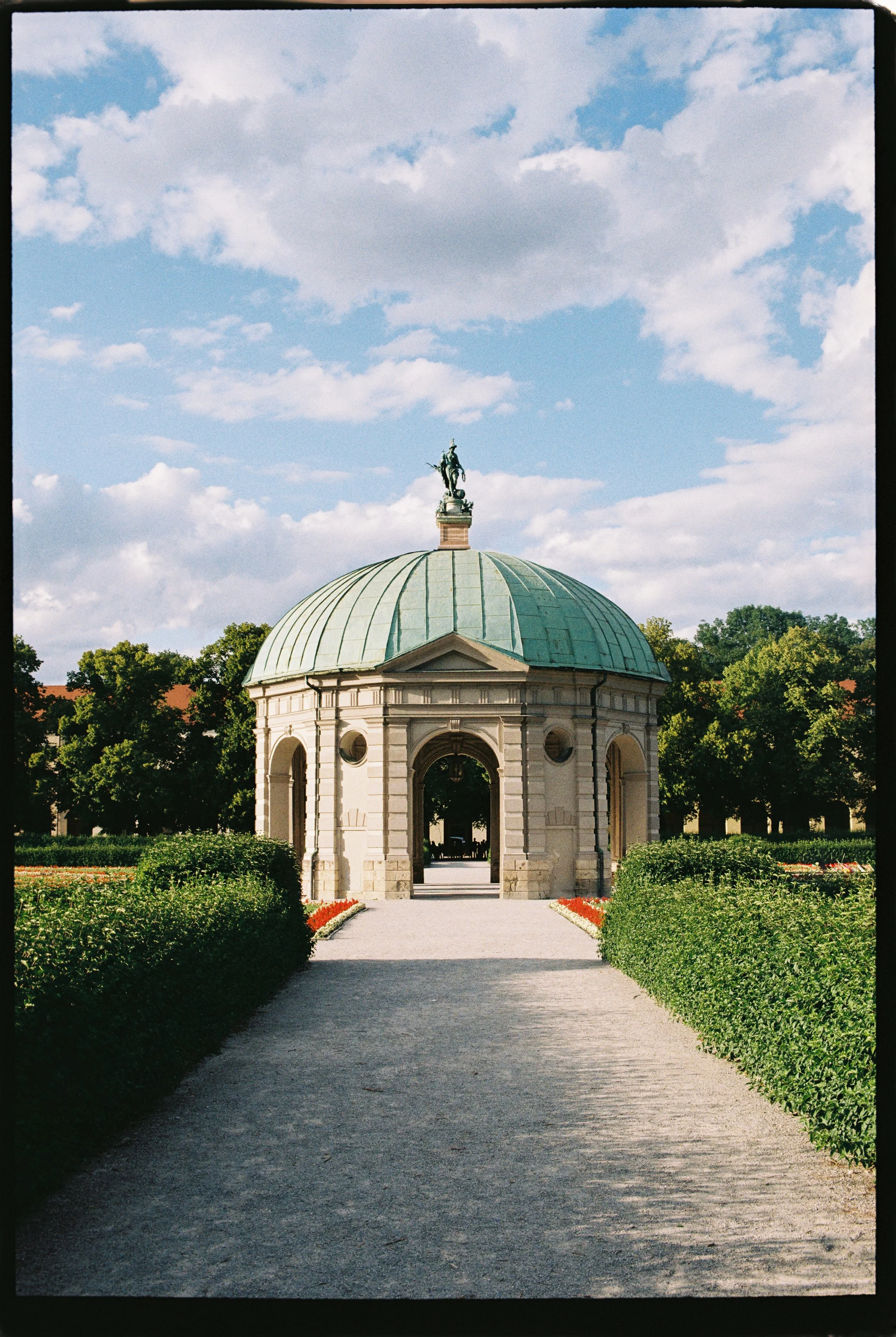 A small, classical-style pavilion with a green-domed roof and arched openings, topped by a statue of a mounted figure, set in a park with garden beds and trees in the background under a partly cloudy sky.