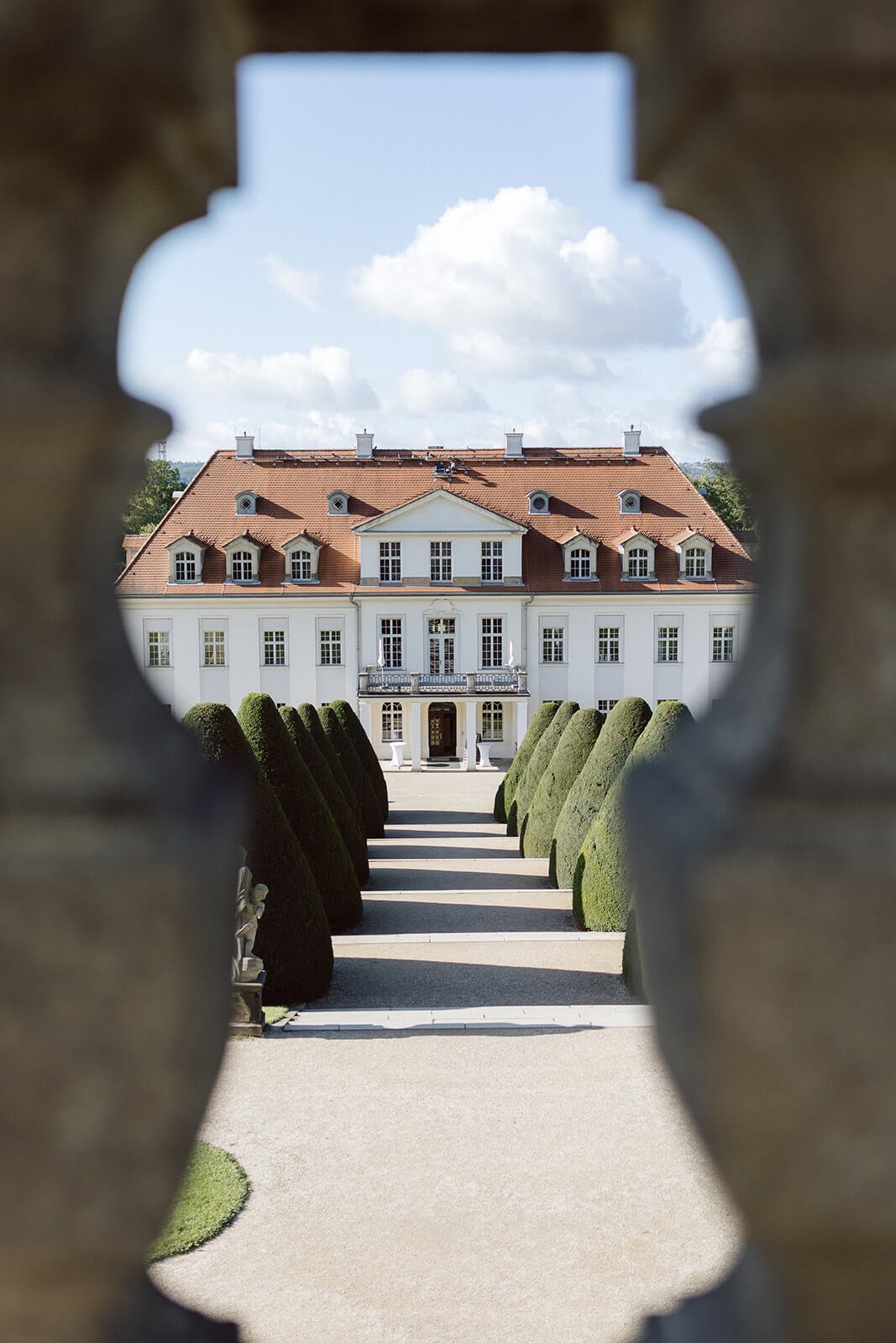 View of a large white mansion with a red roof, framed through a decorative stone opening, with green bushes lining a pathway leading to the entrance.