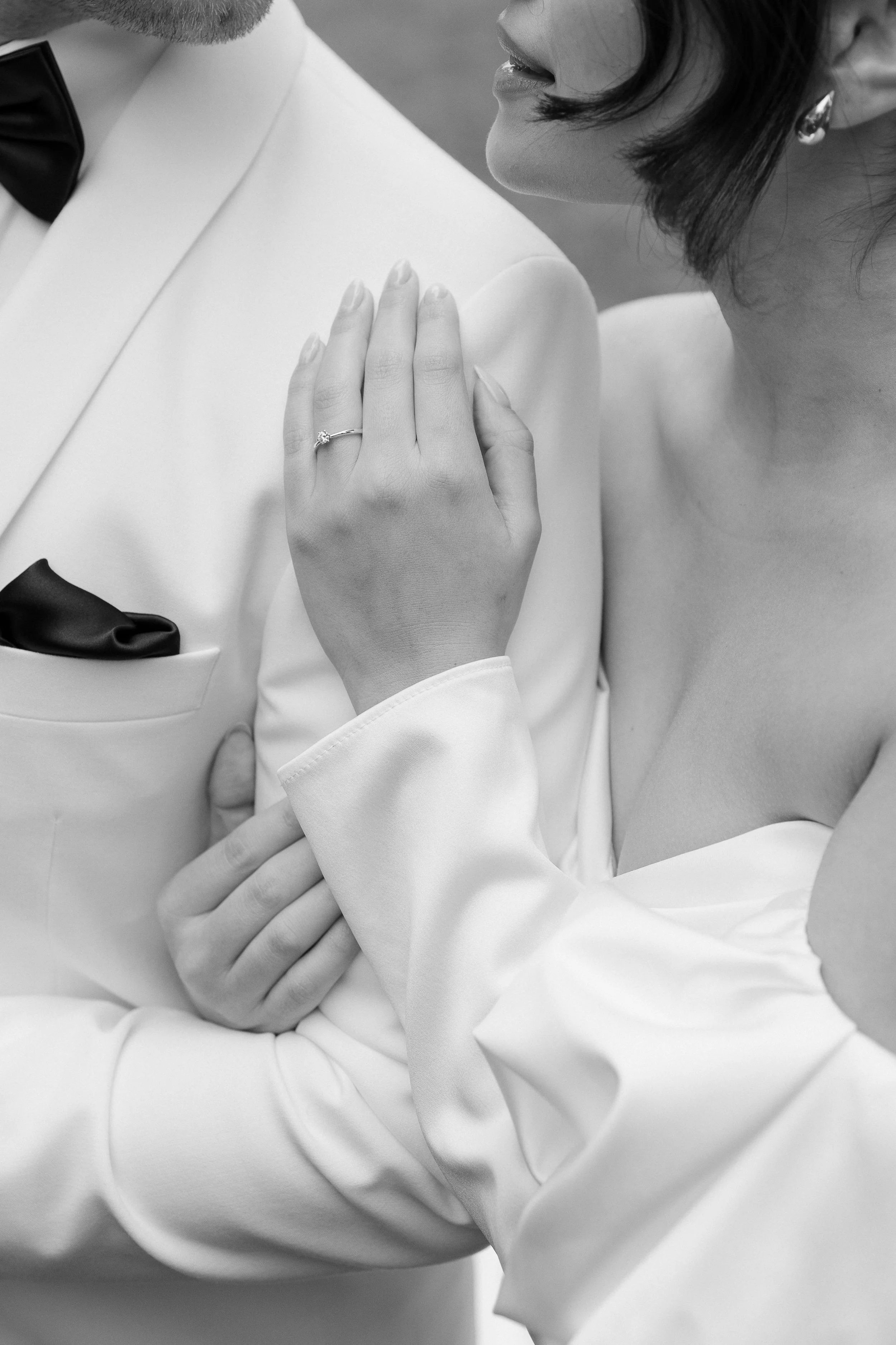 Close-up of a woman with dark hair wearing earrings and a strapless dress, with her hand on her shoulder, showing an engagement ring. A man in a tuxedo stands next to her, with his arm around her. The image is in black and white.