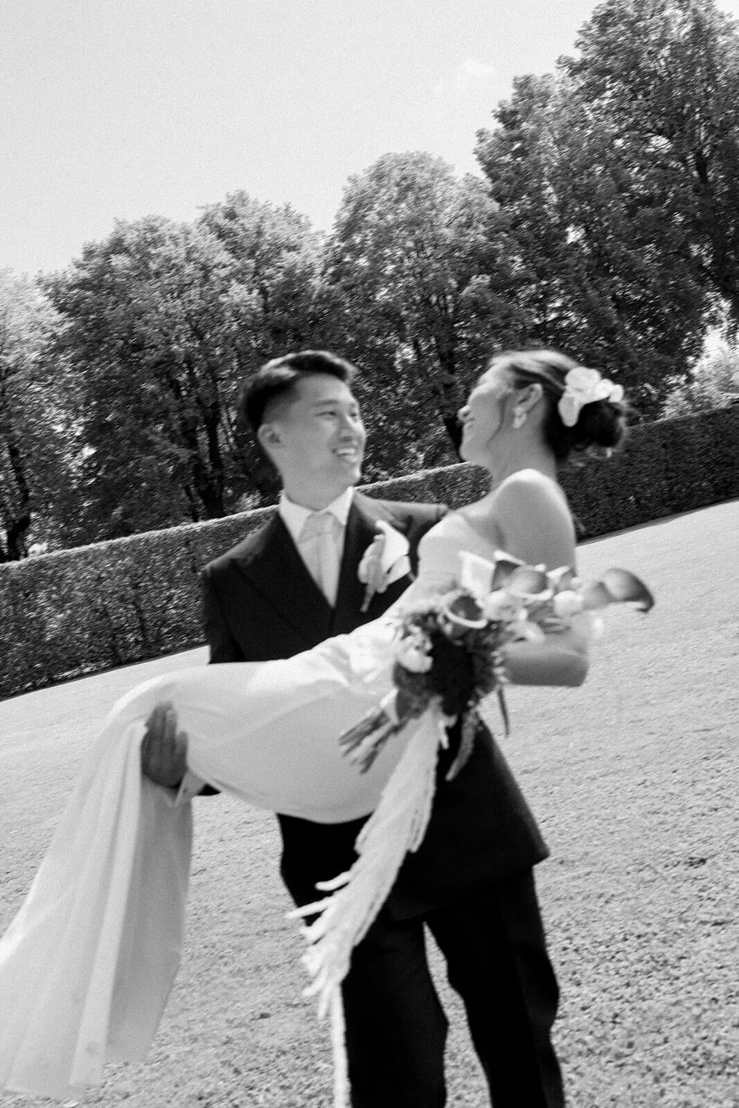 A black-and-white photo of a bride and groom dancing outdoors. The groom is smiling at the bride as she holds a bouquet, and they are surrounded by trees.