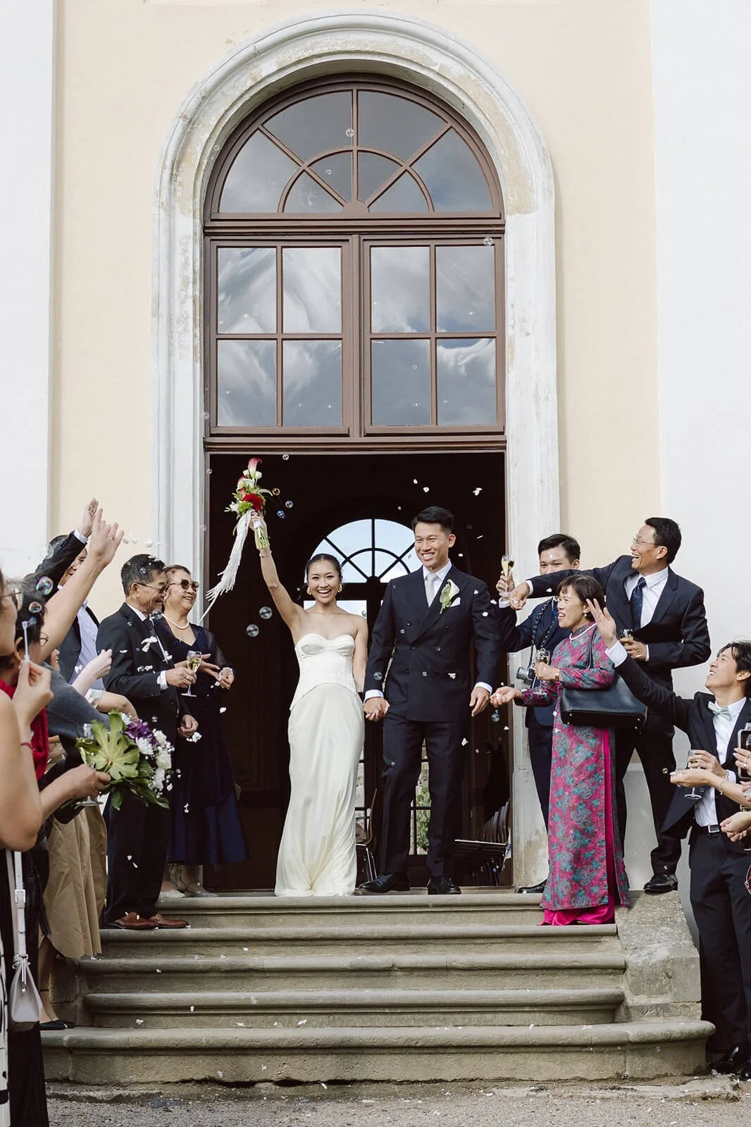 A bride and groom standing on the steps of a church, surrounded by friends and family celebrating with champagne and bubbles.