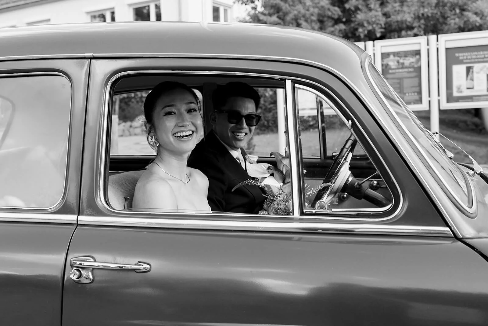 A black and white photo of a smiling woman and man sitting in a vintage car, possibly on their wedding day, with the woman holding a bouquet.