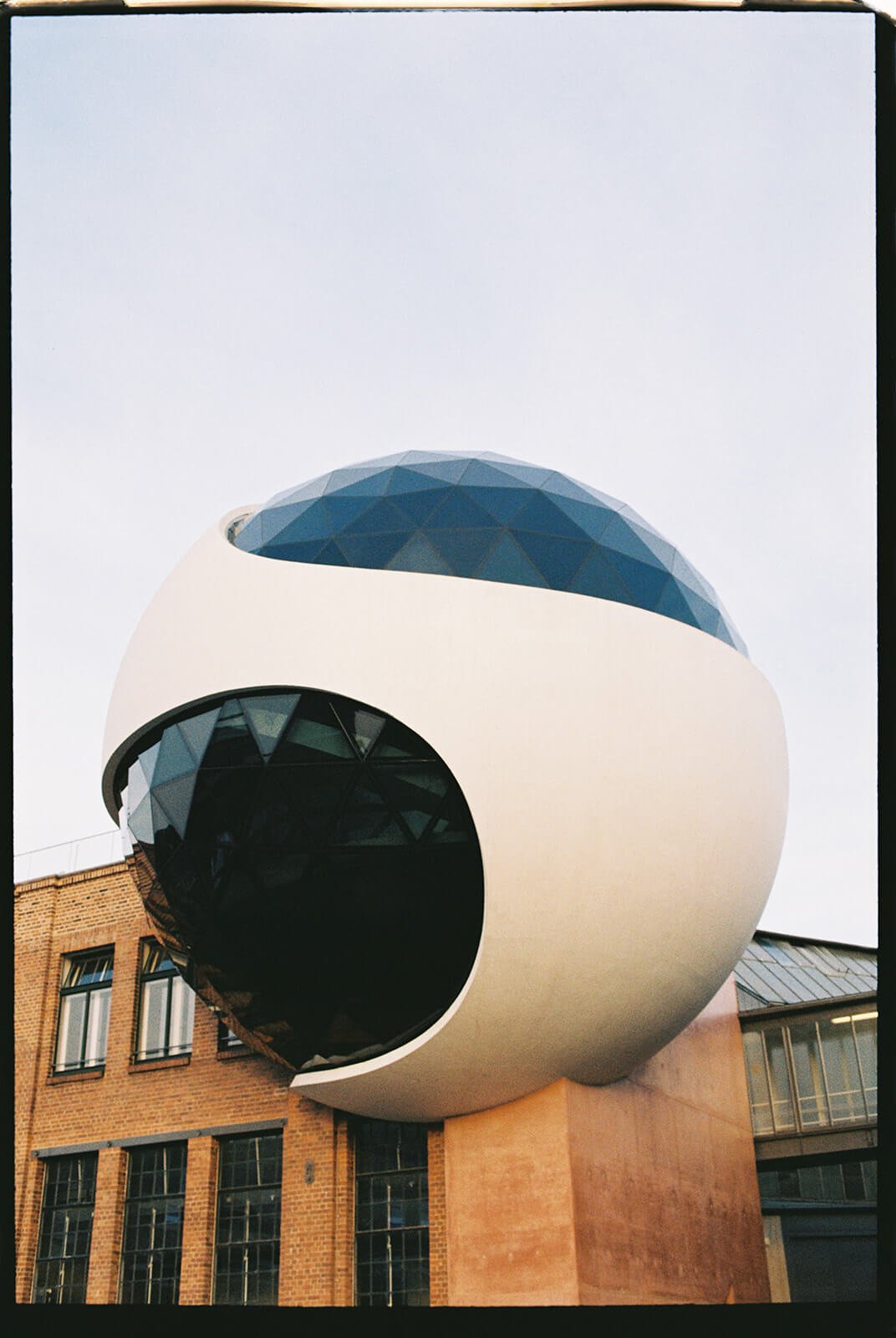 The image shows a modern, spherical building with a large black circular opening and a blue glass dome on top, situated in front of brick buildings.