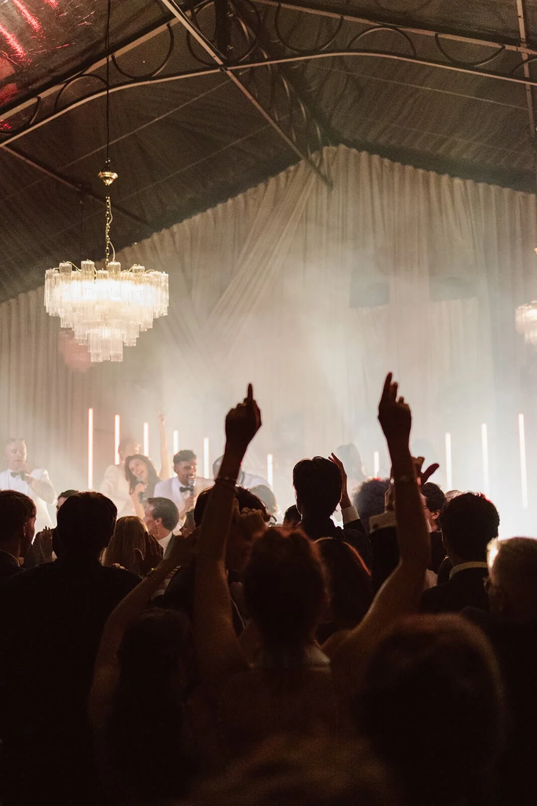 People dancing and celebrating at a formal event with a chandelier and stage lighting in the background.