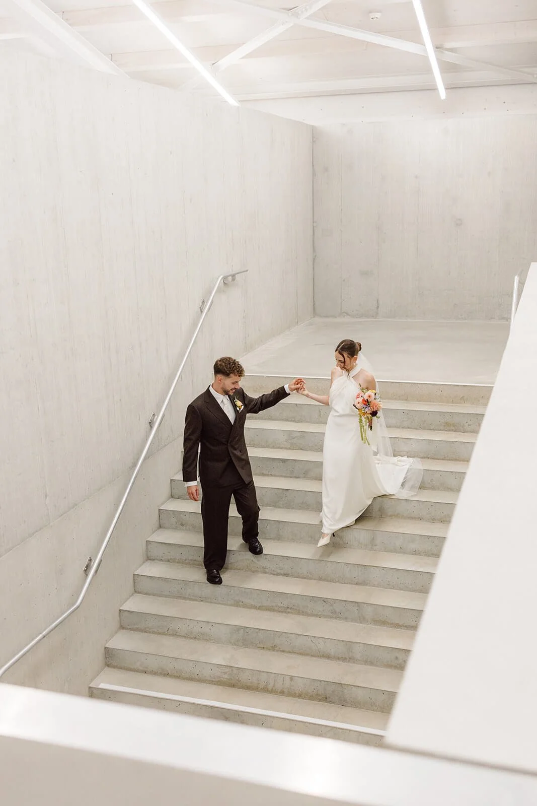 A bride and groom holding hands on a staircase, with the bride in a white wedding dress holding a bouquet of flowers, and the groom in a black suit and tie, in a modern, minimalist interior setting.