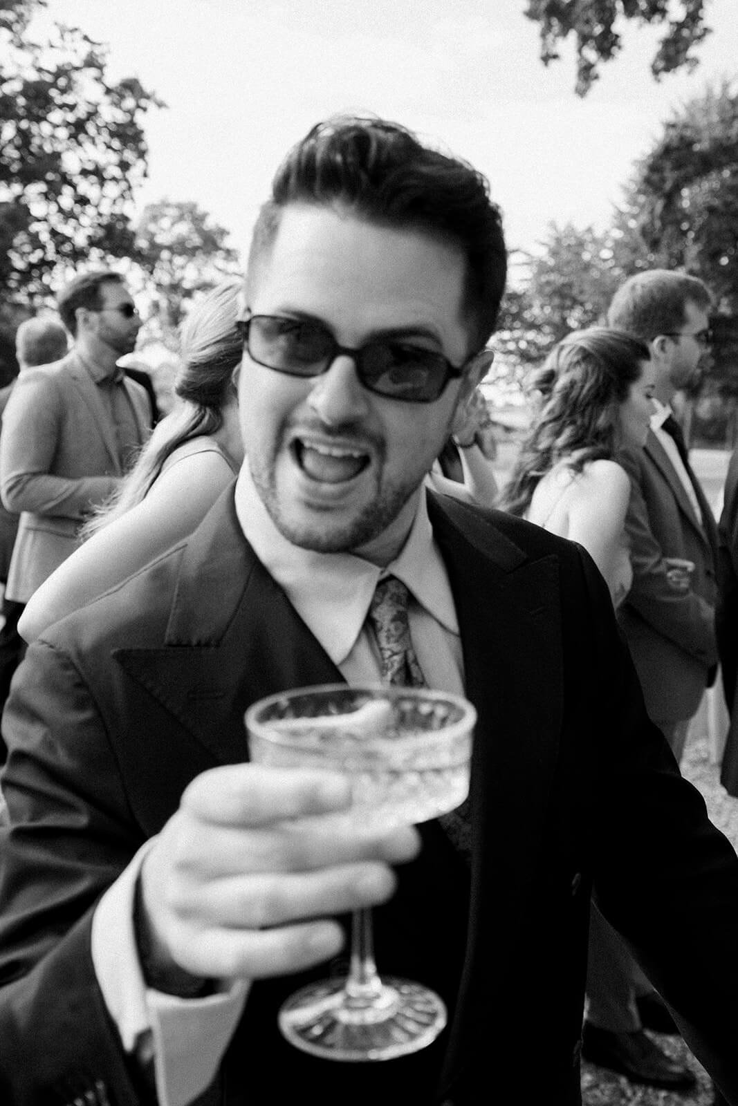 A man in a suit and sunglasses is smiling and holding a glass of wine at an outdoor event with other people dressed in formal attire in the background.