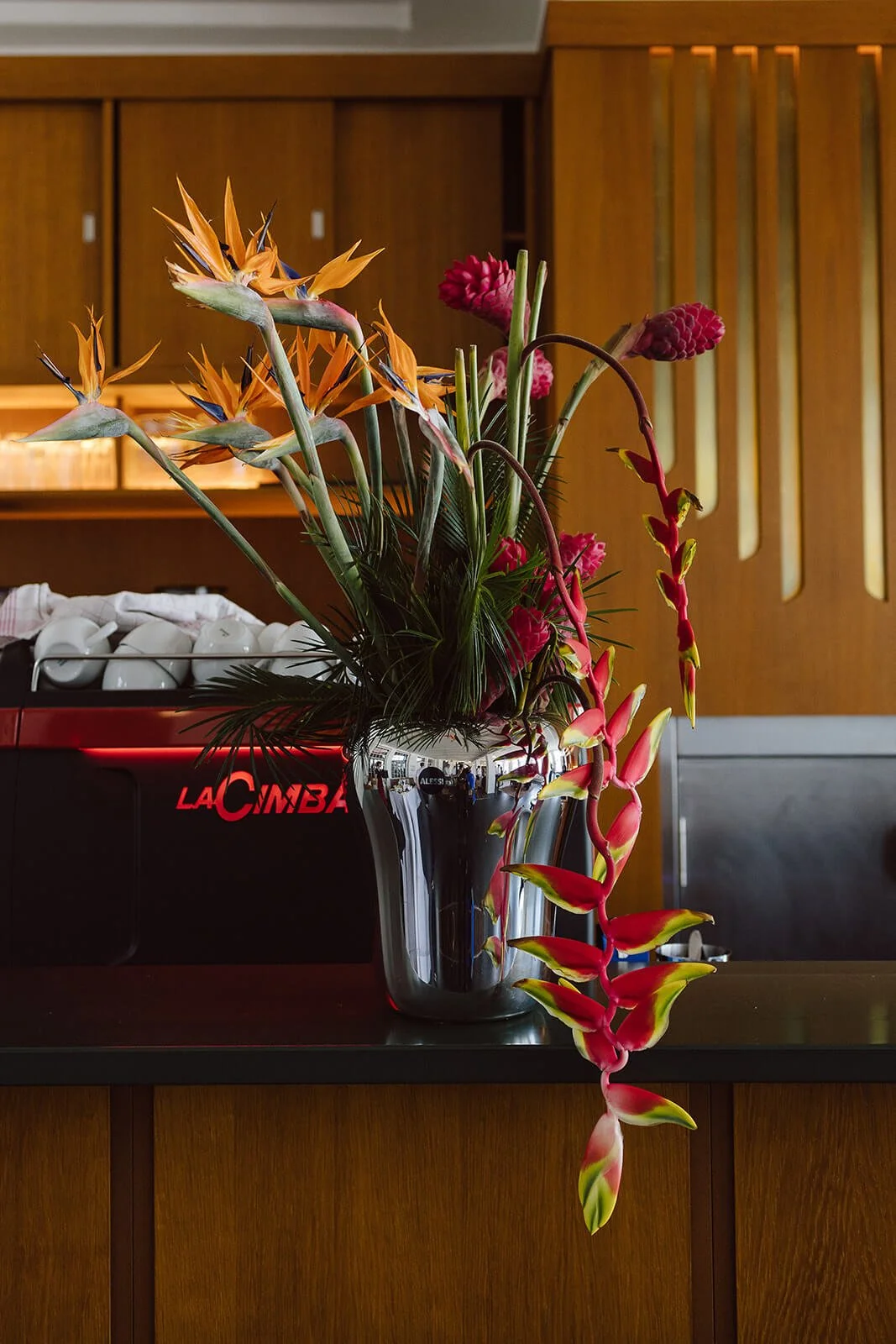 A floral arrangement with orange bird of paradise, pink ginger, and red flowers in a shiny silver vase on a dark countertop inside a room with wooden wall panels.