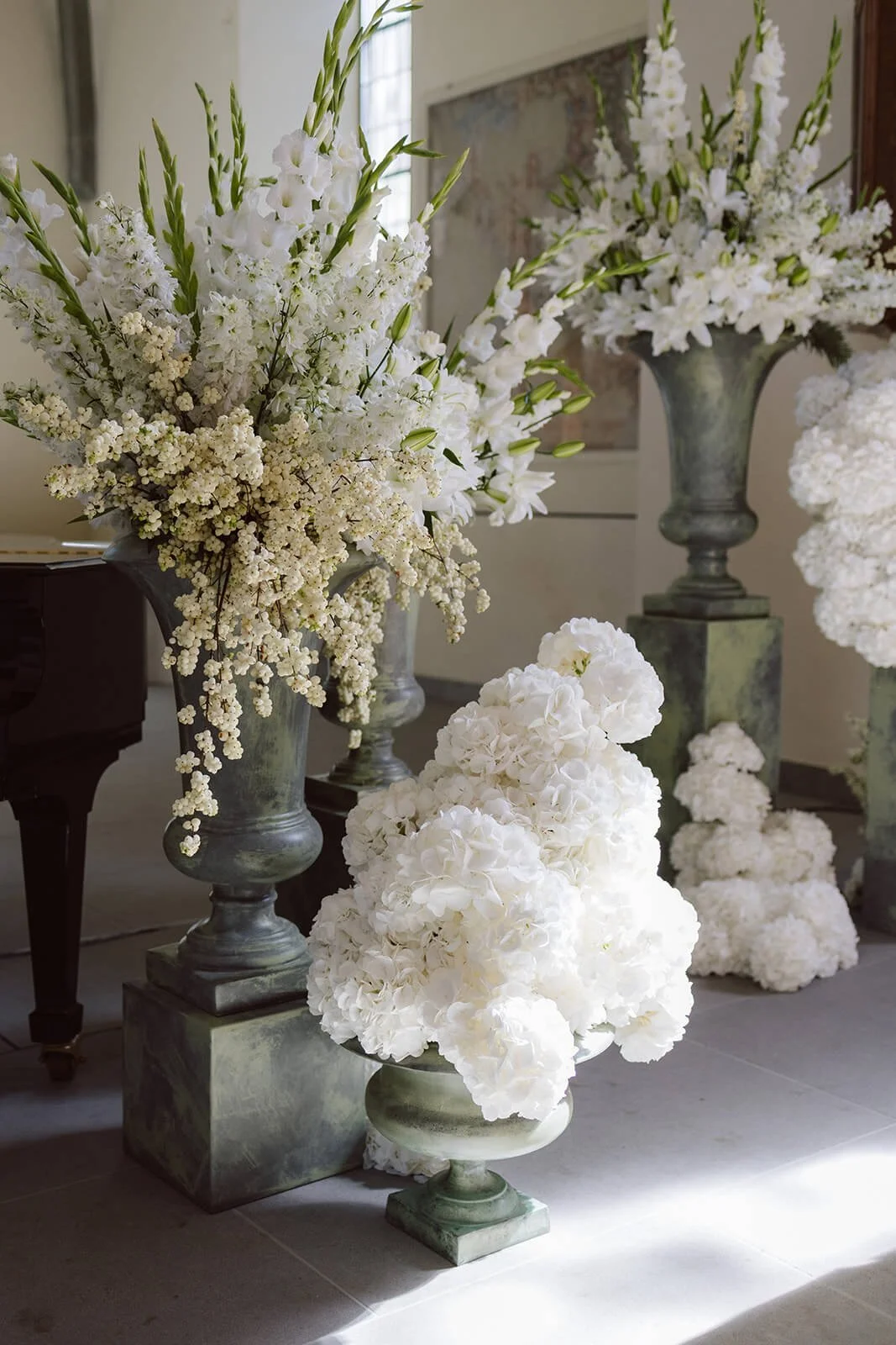 White floral arrangements in tall and short vases, including lilies, hydrangeas, and other white flowers, displayed indoors on a tiled floor.