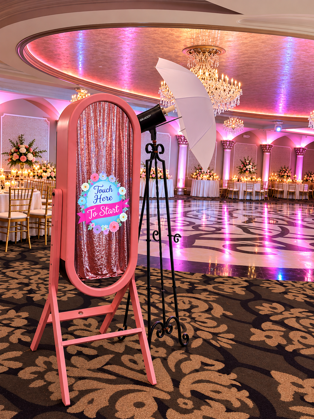 Pink mirror photo booth with umbrella lighting beside a ballroom dance floor, surrounded by crystal chandeliers and pink uplighting.