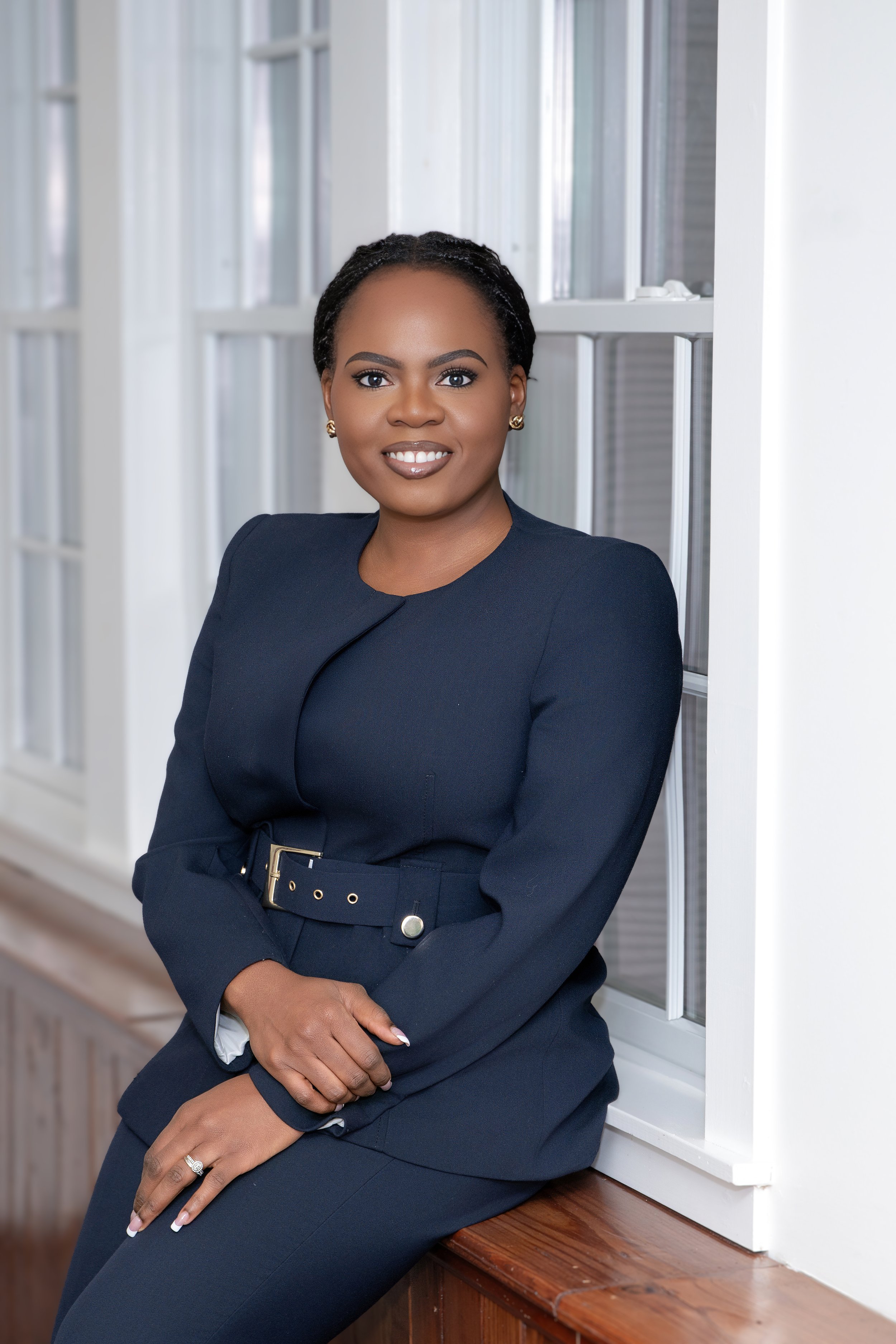 A woman with short, curly black hair wearing a navy blue blazer and matching pants, sitting on a wooden bench by a large window, smiling at the camera.