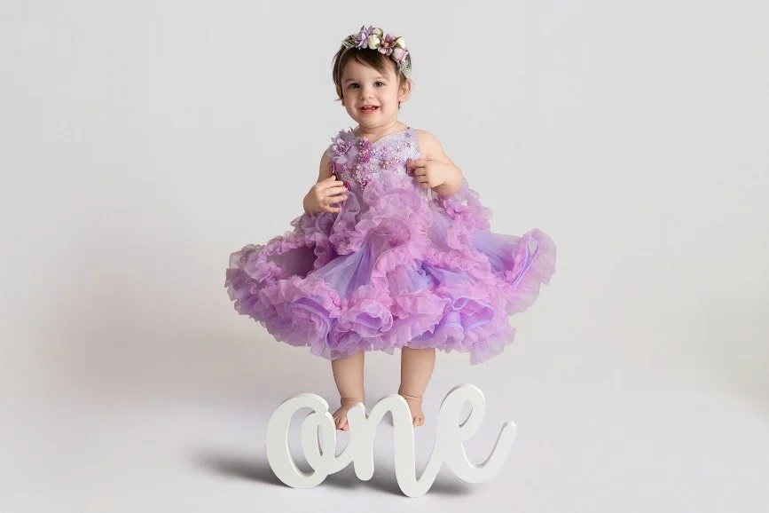 One year old baby girl in purple ruffle dress standing over “one” sign during first birthday studio photoshoot in Mercer County NJ