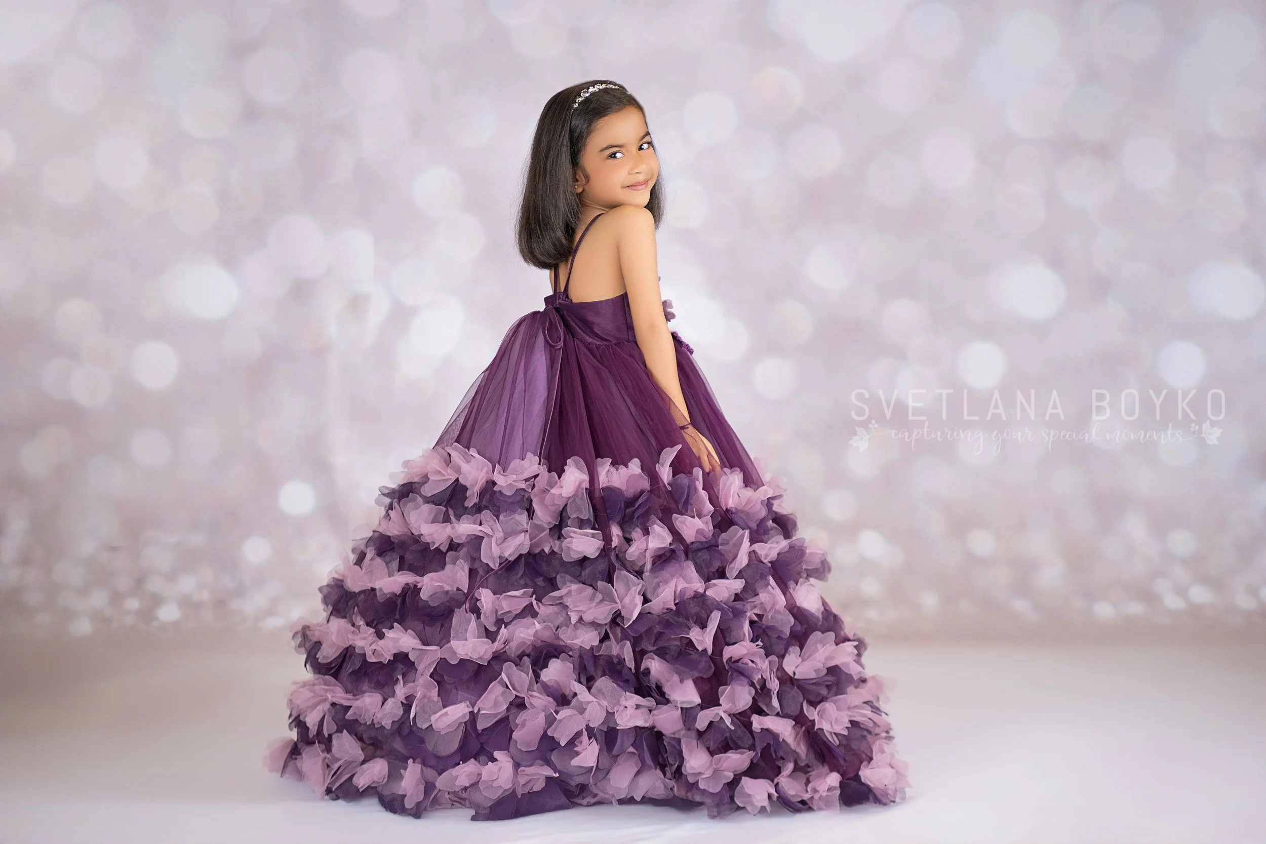 Young girl in a purple gown with a layered tulle skirt, standing against a sparkling, bokeh background.
