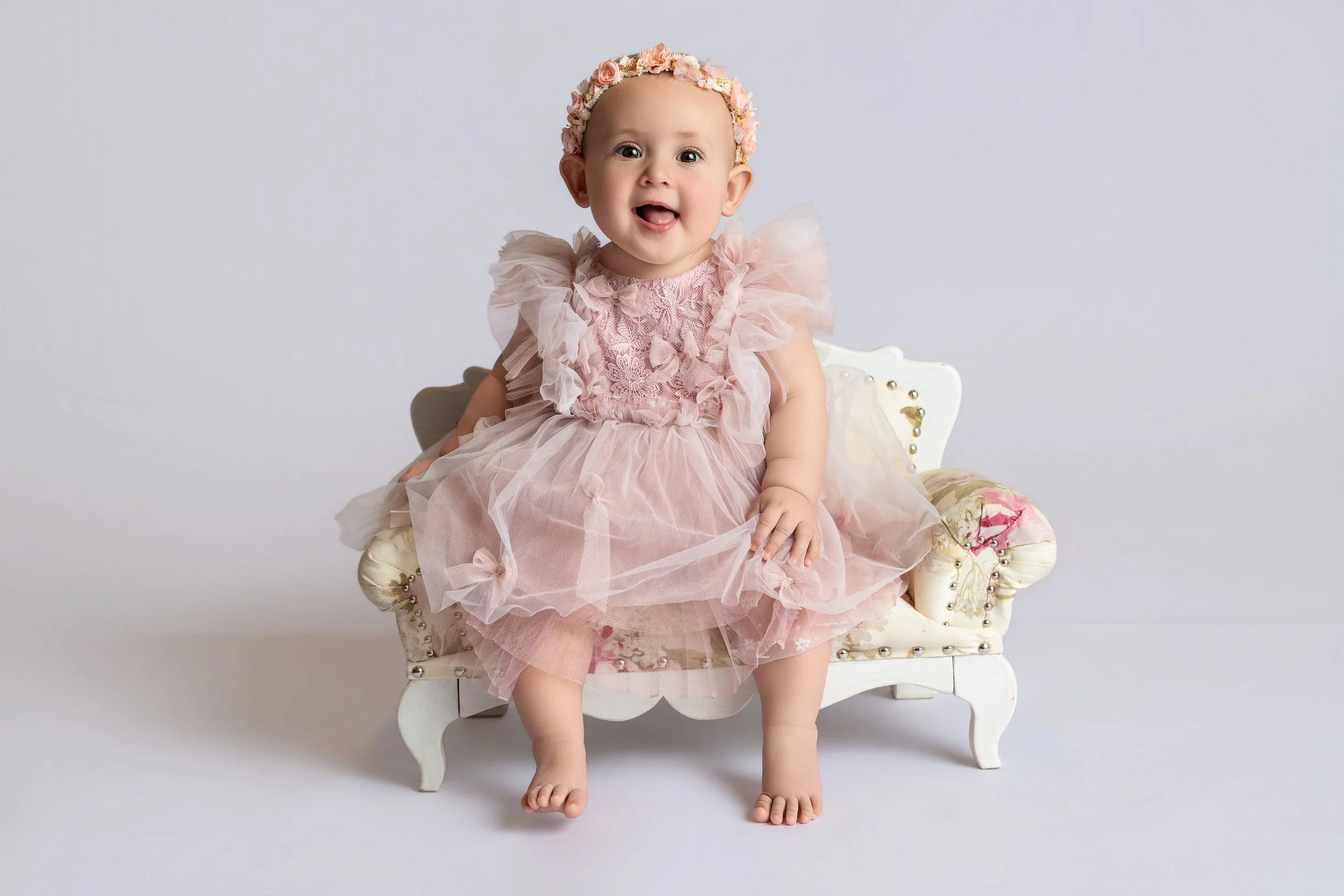 A baby girl wearing a pink dress and a flower headband, sitting on a small vintage-style white bench with pink and gold upholstery, smiling at the camera against a plain light-colored background.