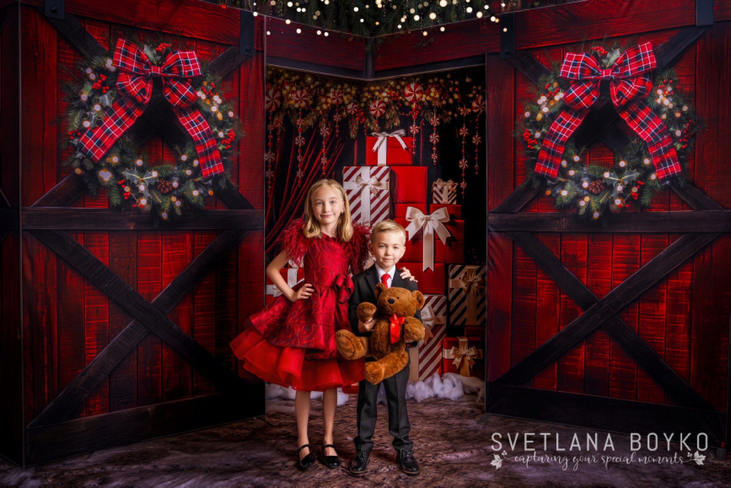 Children posing in a festive red Christmas barn set with glowing lights, holiday wreaths, wrapped presents, and classic seasonal décor during a holiday mini session in New Jersey.