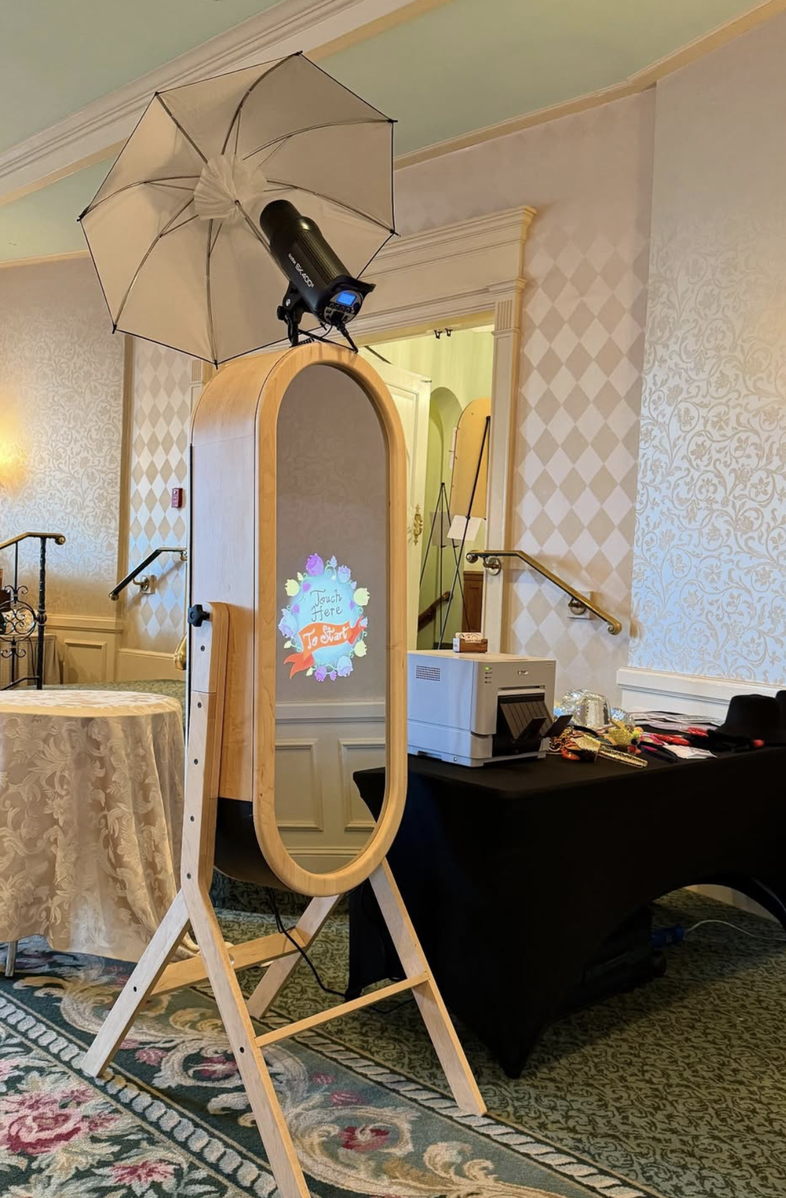 Photo of a room set up for a photo booth, with a wooden full-length mirror, a black table with a printer and other items, a large umbrella with a strobe light mounted on top, and a decorative sign reading 'Touch & Fête to start,' in a room with patterned wallpaper and a patterned rug.