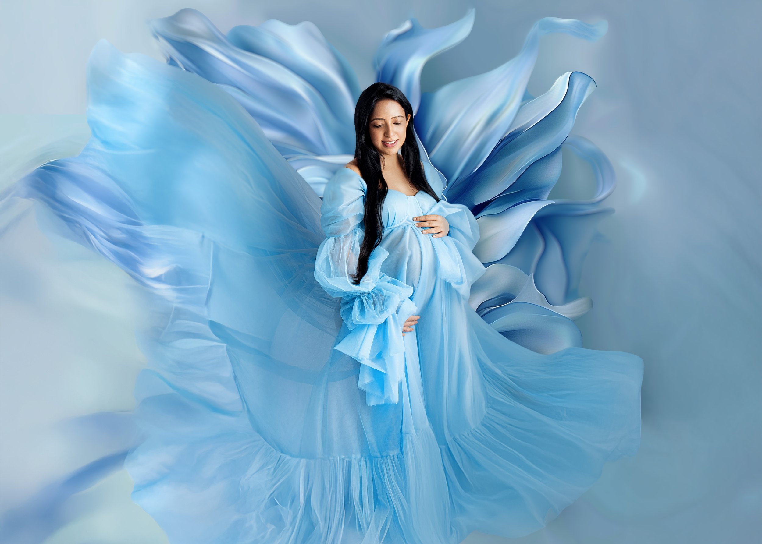 A pregnant woman in a flowing, sheer, blue dress with large, ruffled sleeves, standing in front of a background designed to look like a large, blue flower with layered petals.