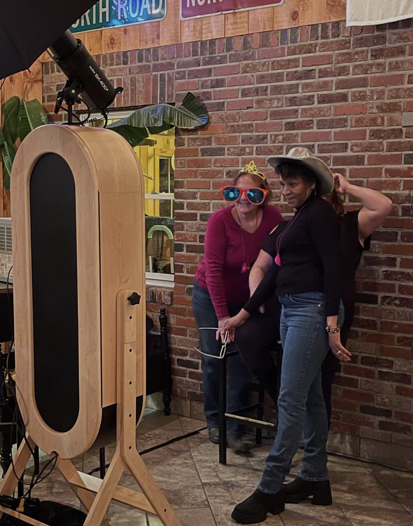 Two women are taking a photo in front of a brick wall, one wearing a playful pink crown and large red sunglasses, and the other wearing a large wide-brimmed hat. They seem to be enjoying a light-hearted moment at an indoor social gathering or photo booth.
