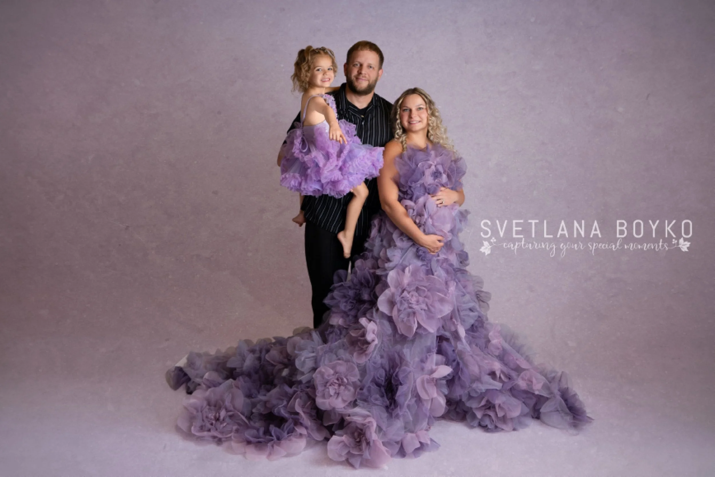 A man and two young girls posing together in front of a plain gray background. The girls are dressed in purple tulle dresses with floral details, and the man is wearing a black striped shirt.