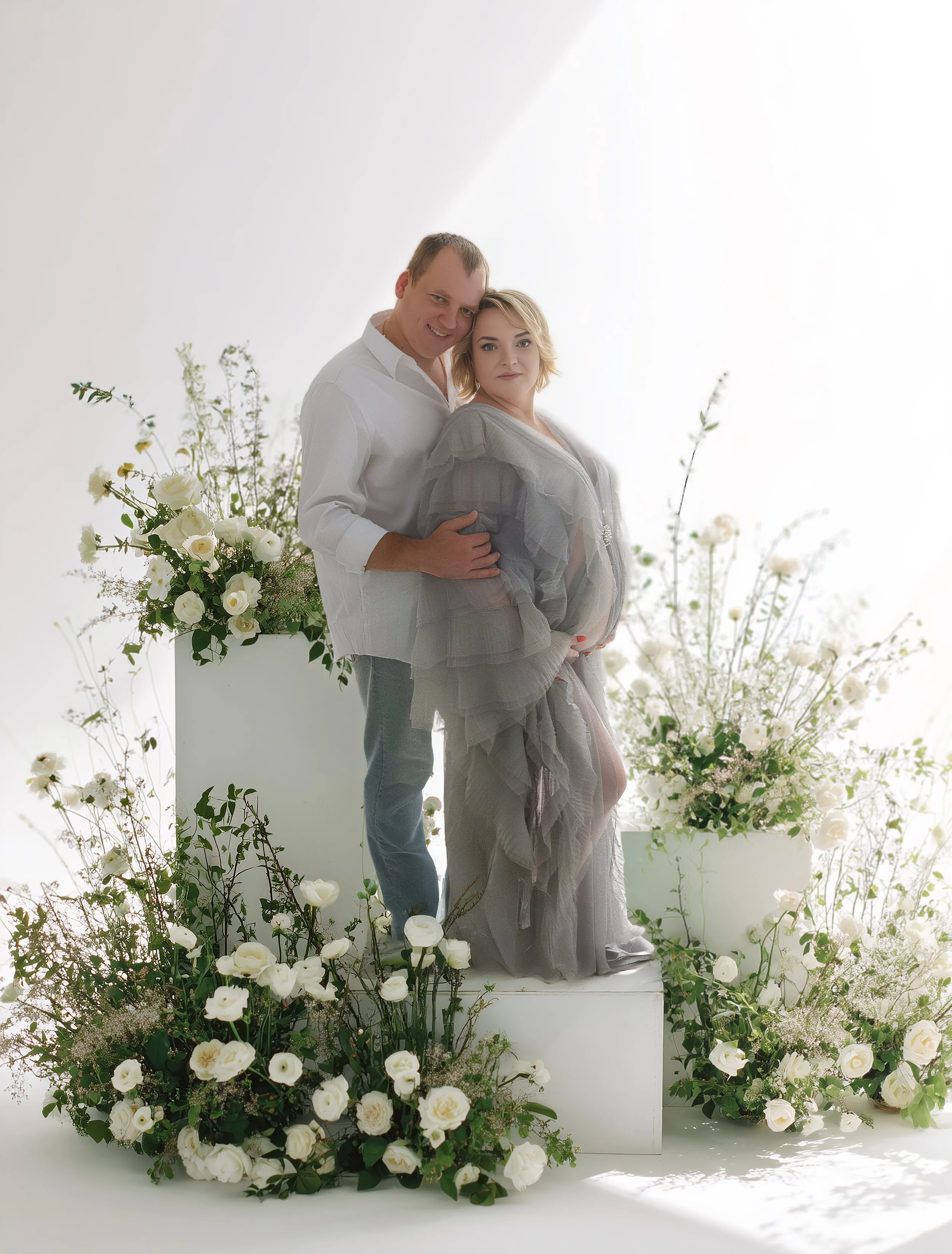 A couple posing together among white flowers and greenery in a bright studio setting.