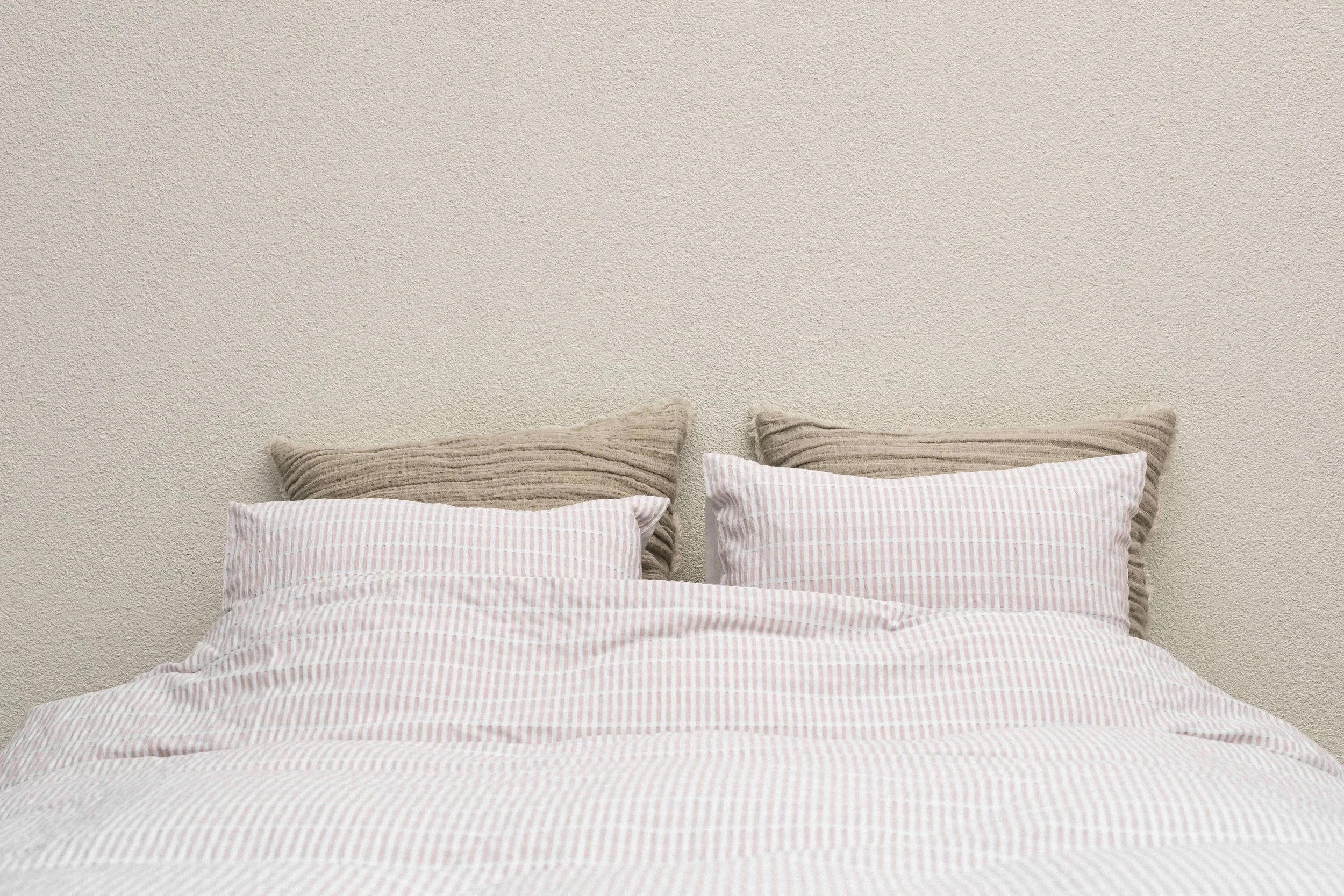 A neatly made bed with two light pink and white striped pillows and two beige textured pillows against a plain, textured off-white wall.