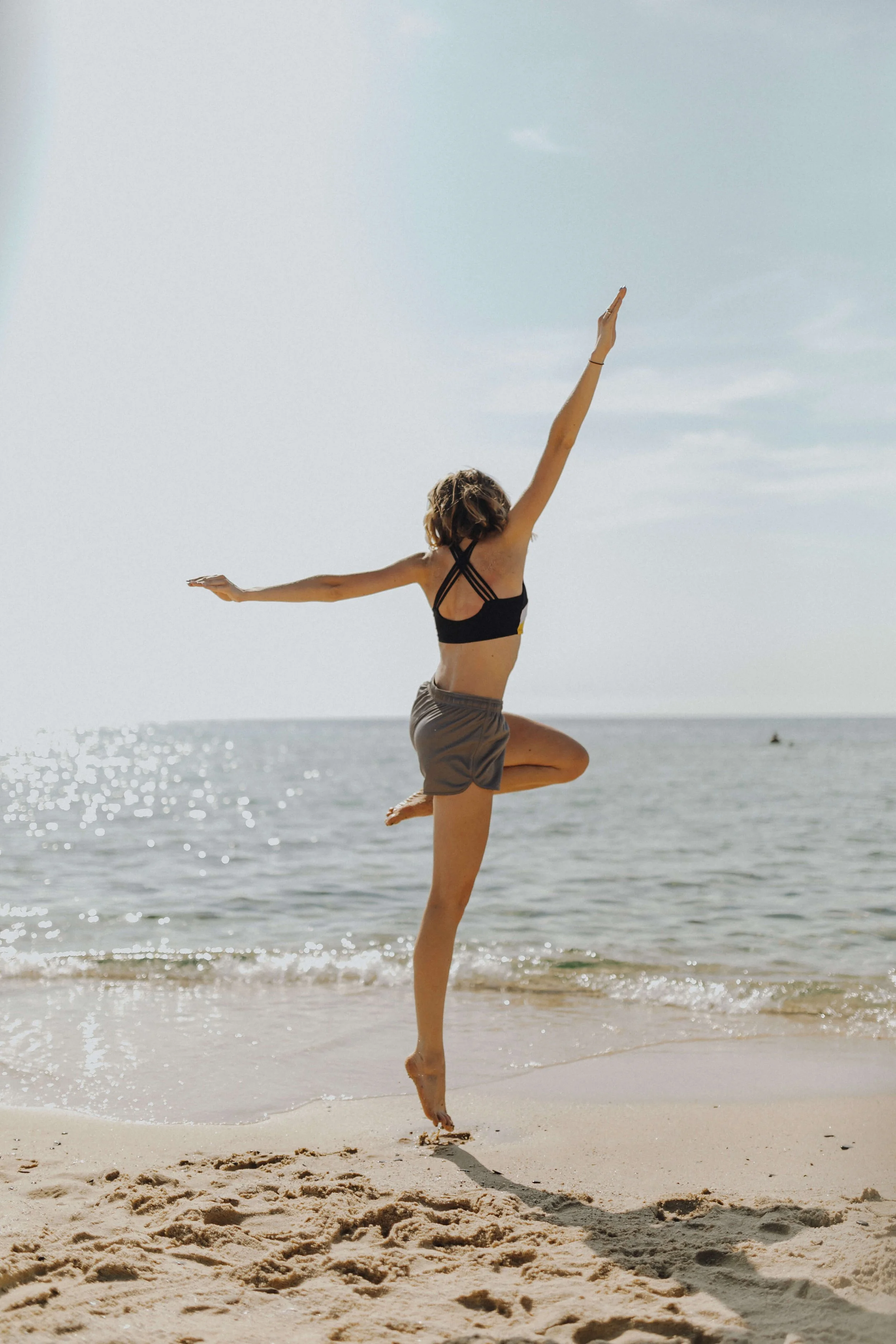 A woman practicing yoga on the beach, balancing on one leg with arms outstretched, wearing a sports bra and shorts.
