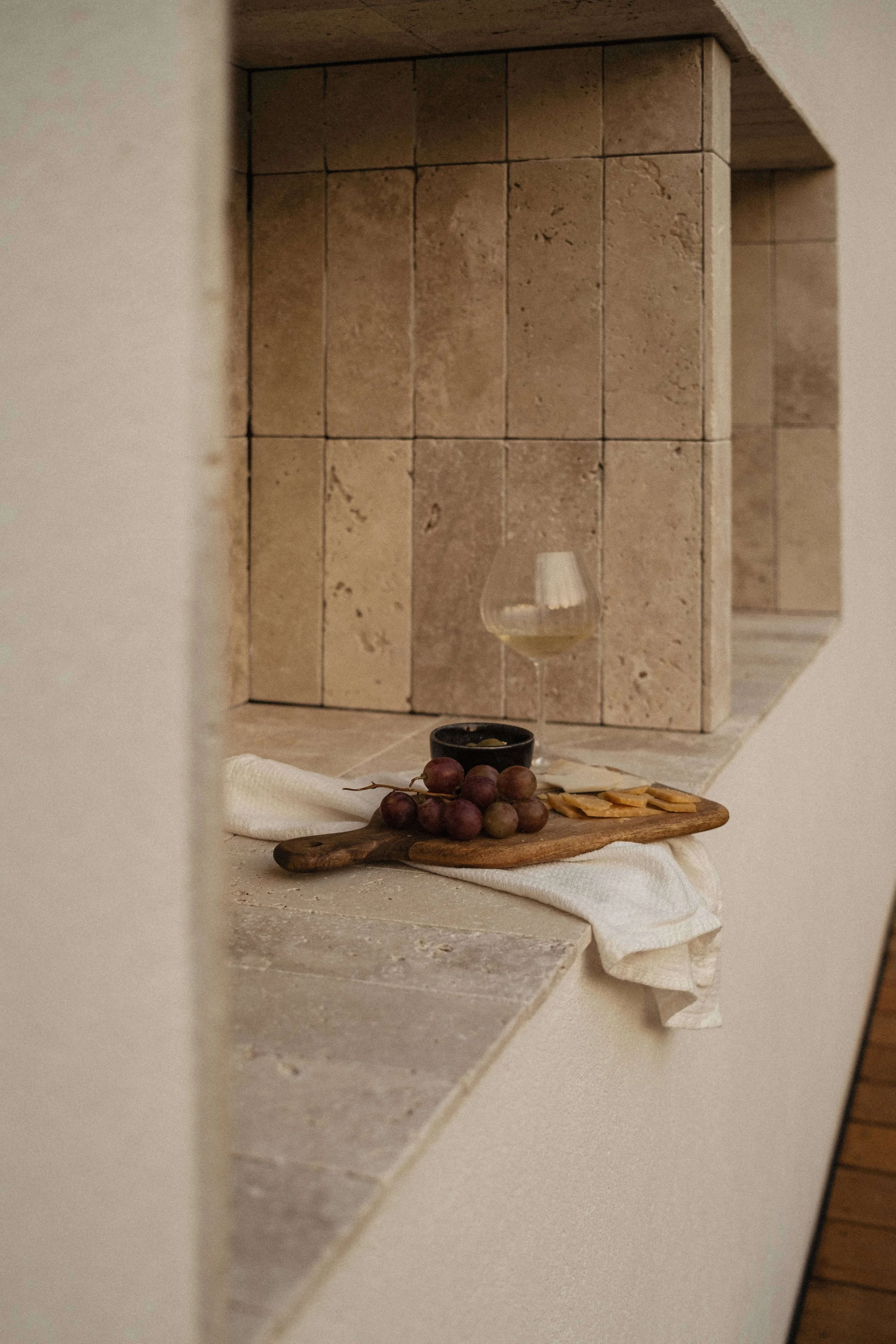 A stone surface with a bunch of grapes, a small black bowl, a glass of white wine, and a cloth napkin; background shows a tiled wall with a small open shelf.