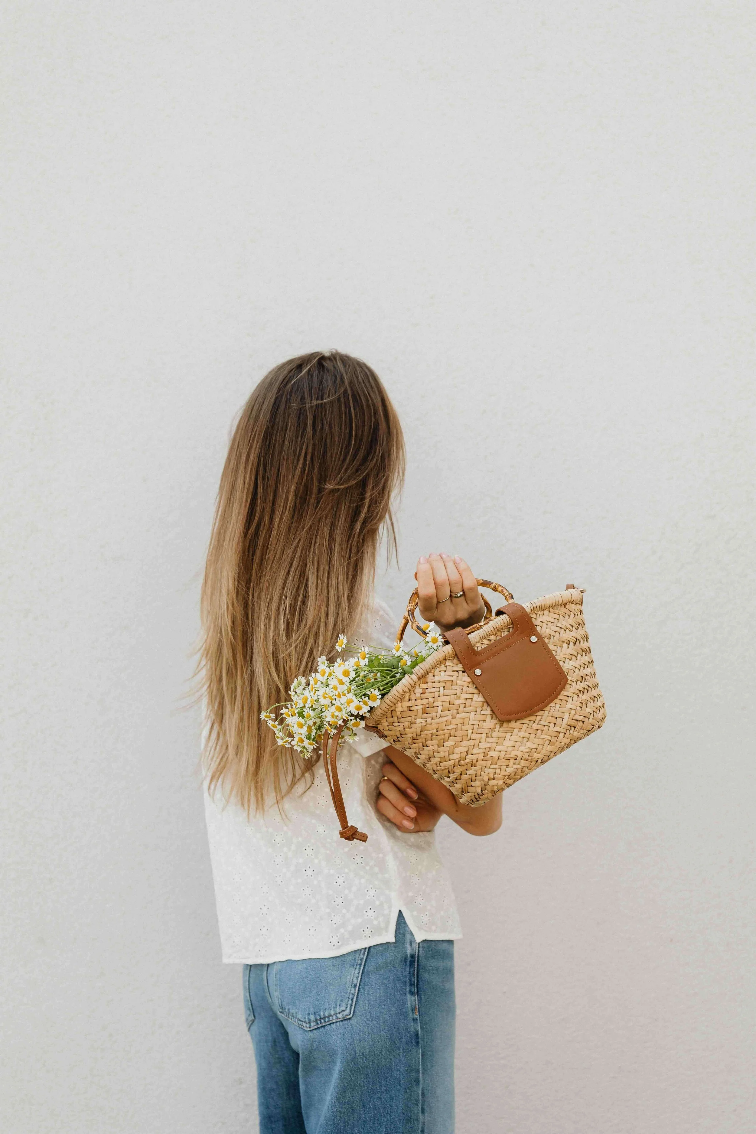 A woman with long, light brown hair wearing a white eyelet blouse and blue jeans, holding a woven straw handbag with daisies inside, against a plain white background.