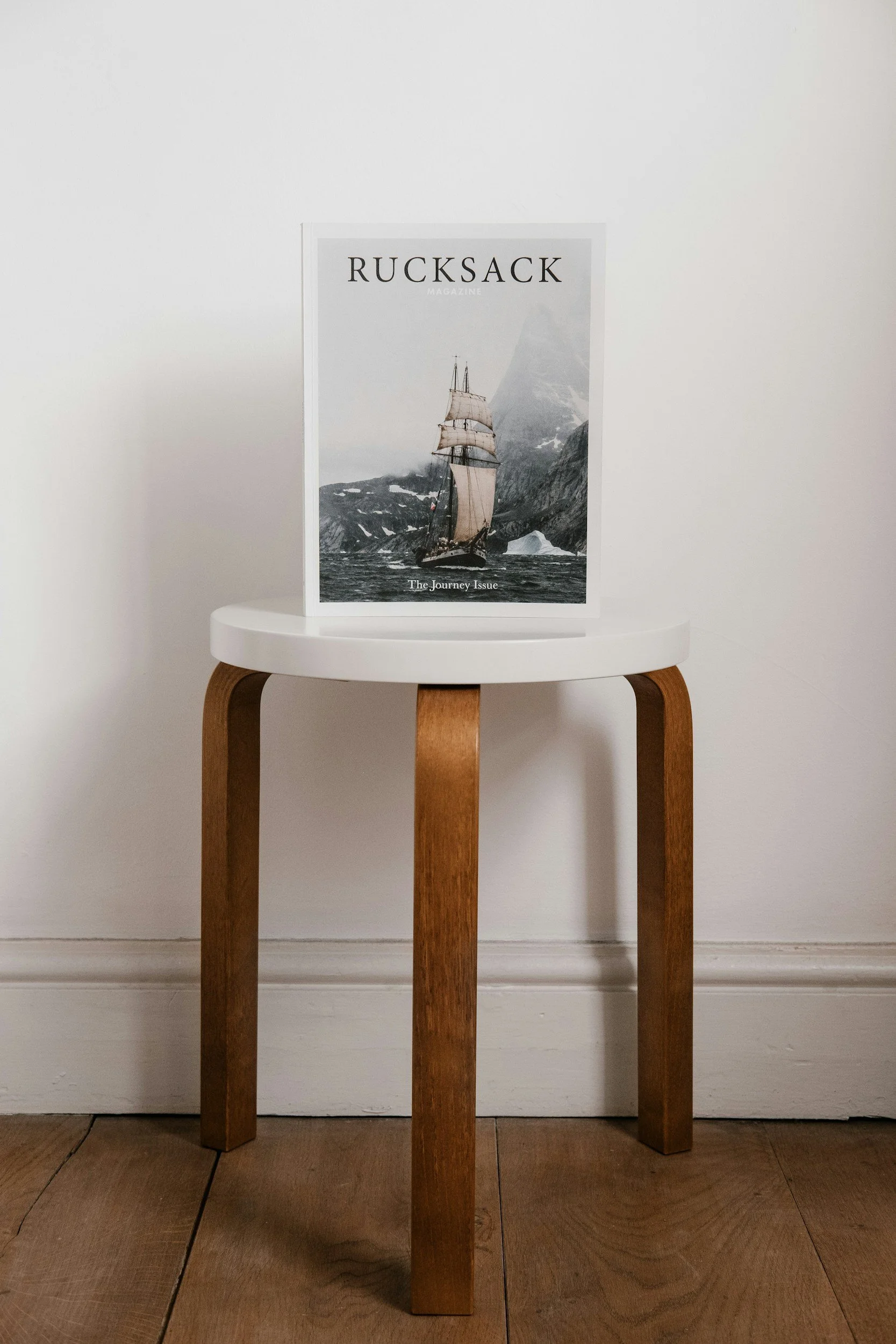 Magazine titled 'Rucksack' displayed on a small wooden stool against a white wall, featuring a sailing ship and snow-covered mountains on the cover.