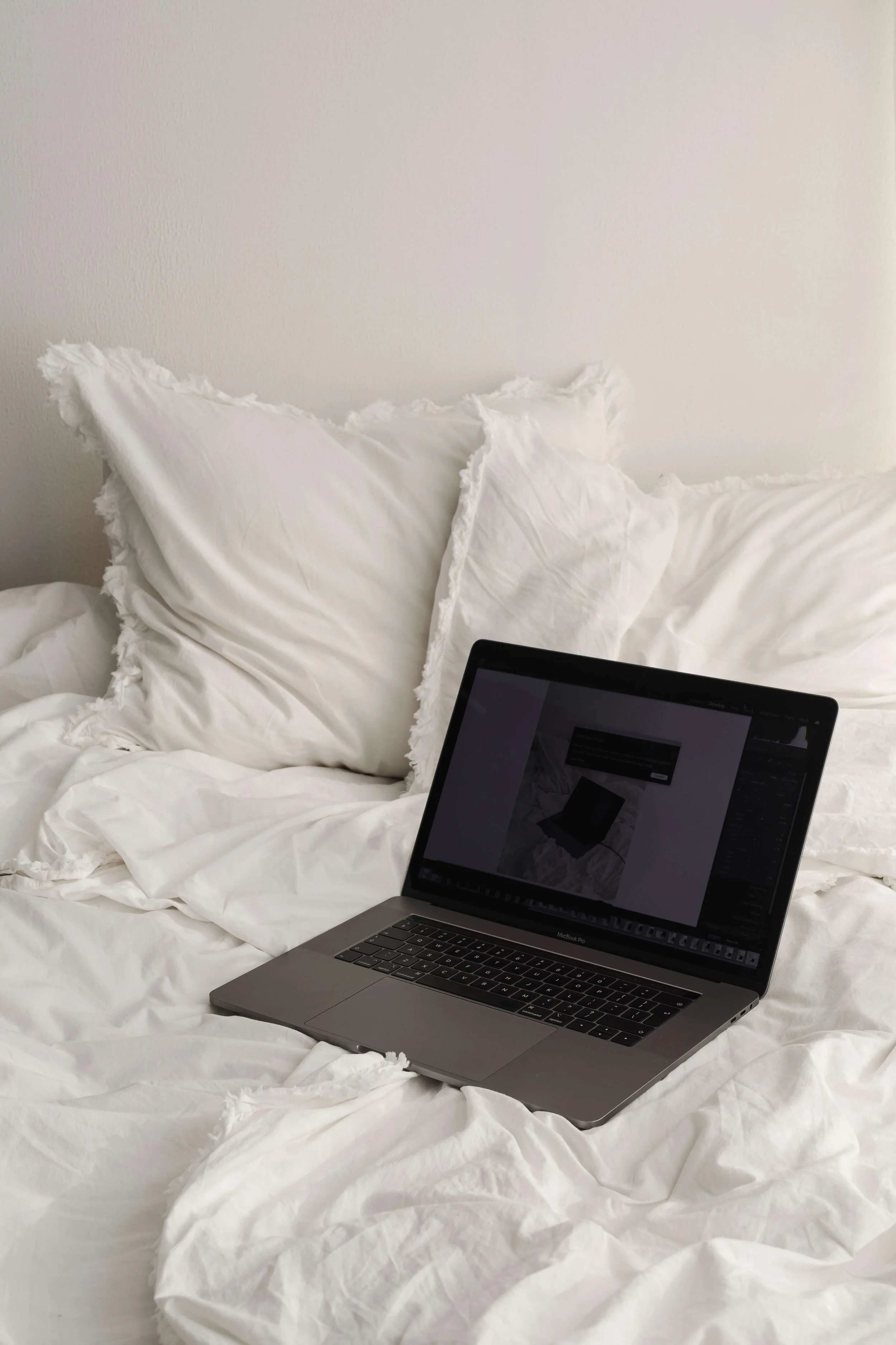 A silver laptop resting on a rumpled white bed with white pillows and a white wall in the background.